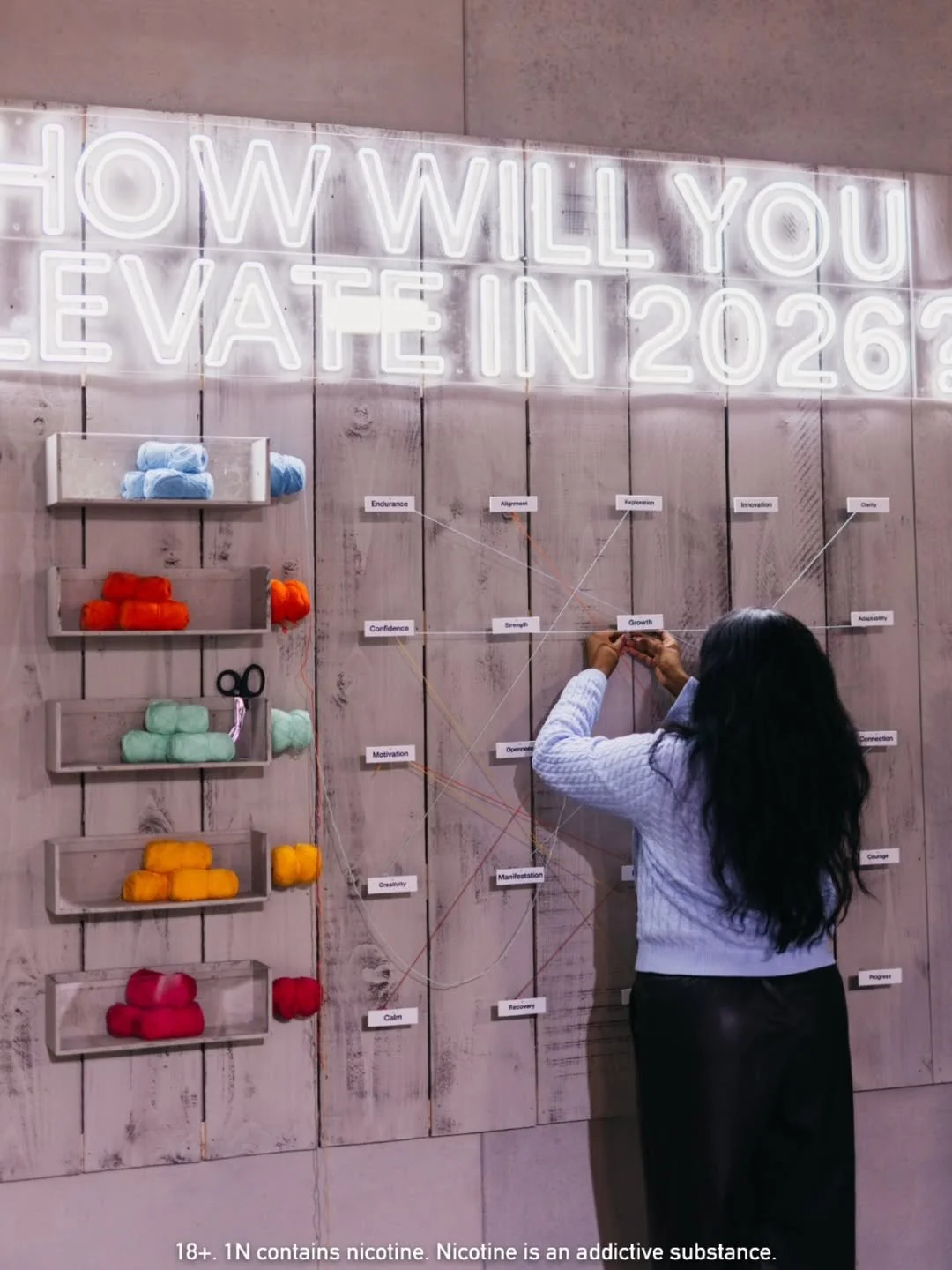 A person with long dark hair in a light-colored sweater and dark pants is working on a visual display board. The board has colorful yarn balls on shelves and a neon sign that reads, "How will you vote in 2026?" A diagram with connected labels such as