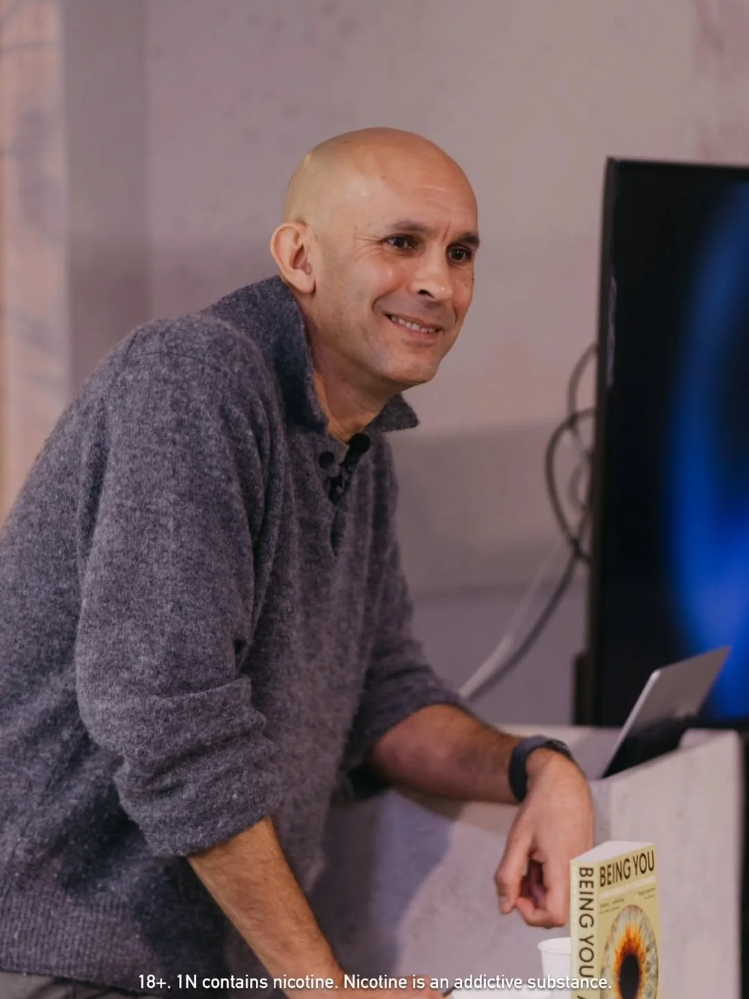 A bald man in a gray sweater smiling while leaning on a counter with a book titled "Being You" on it, next to a computer monitor.
