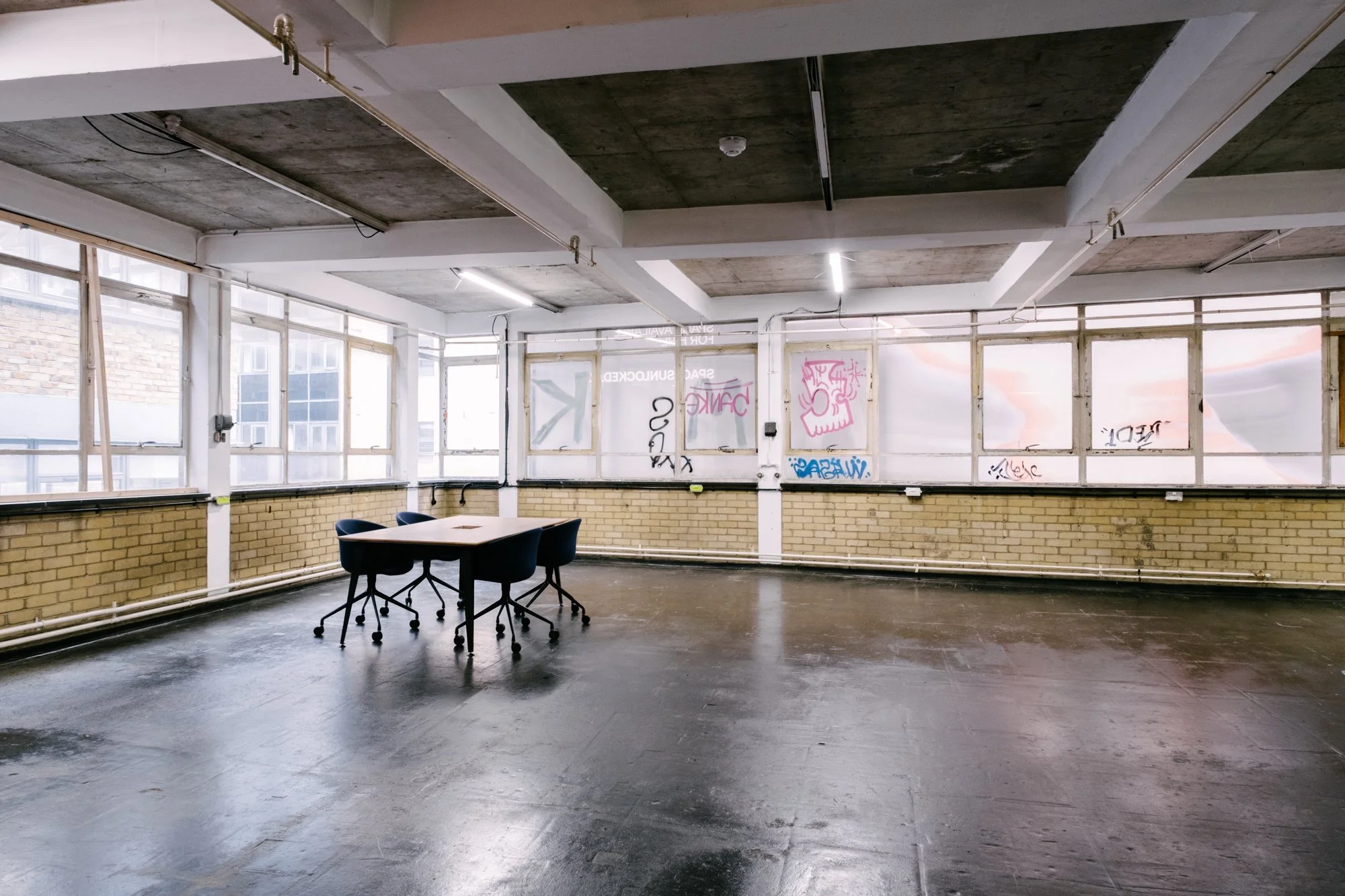 An empty room with large windows, a brick lower wall, and graffiti on the upper wall panels. There are a few black chairs and a table in the center, and the ceiling has exposed concrete with lighting fixtures.