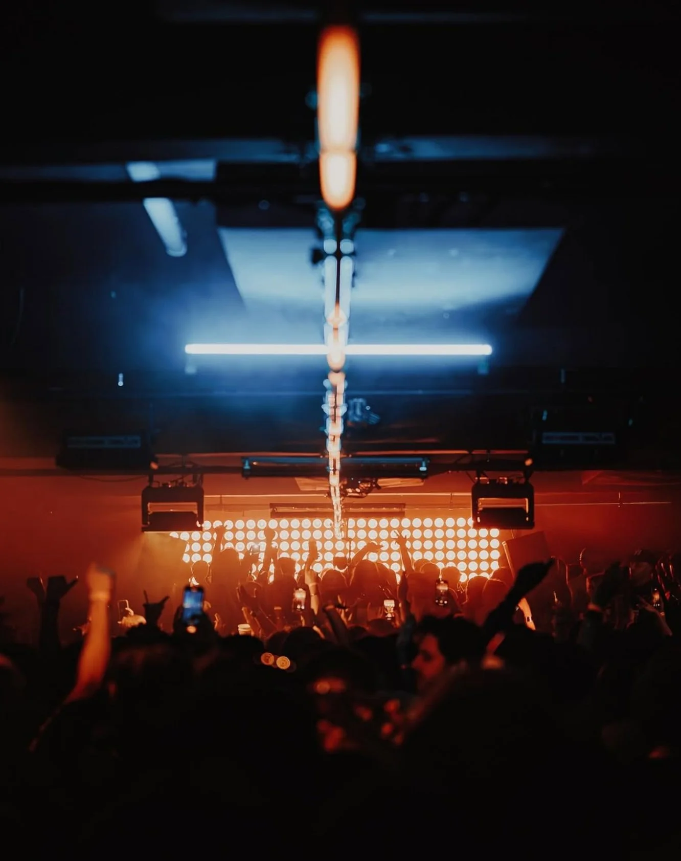 Crowd of people at a concert or nightclub with raised hands, illuminated by colorful stage lights, with a DJ or performer in front of a LED backdrop.