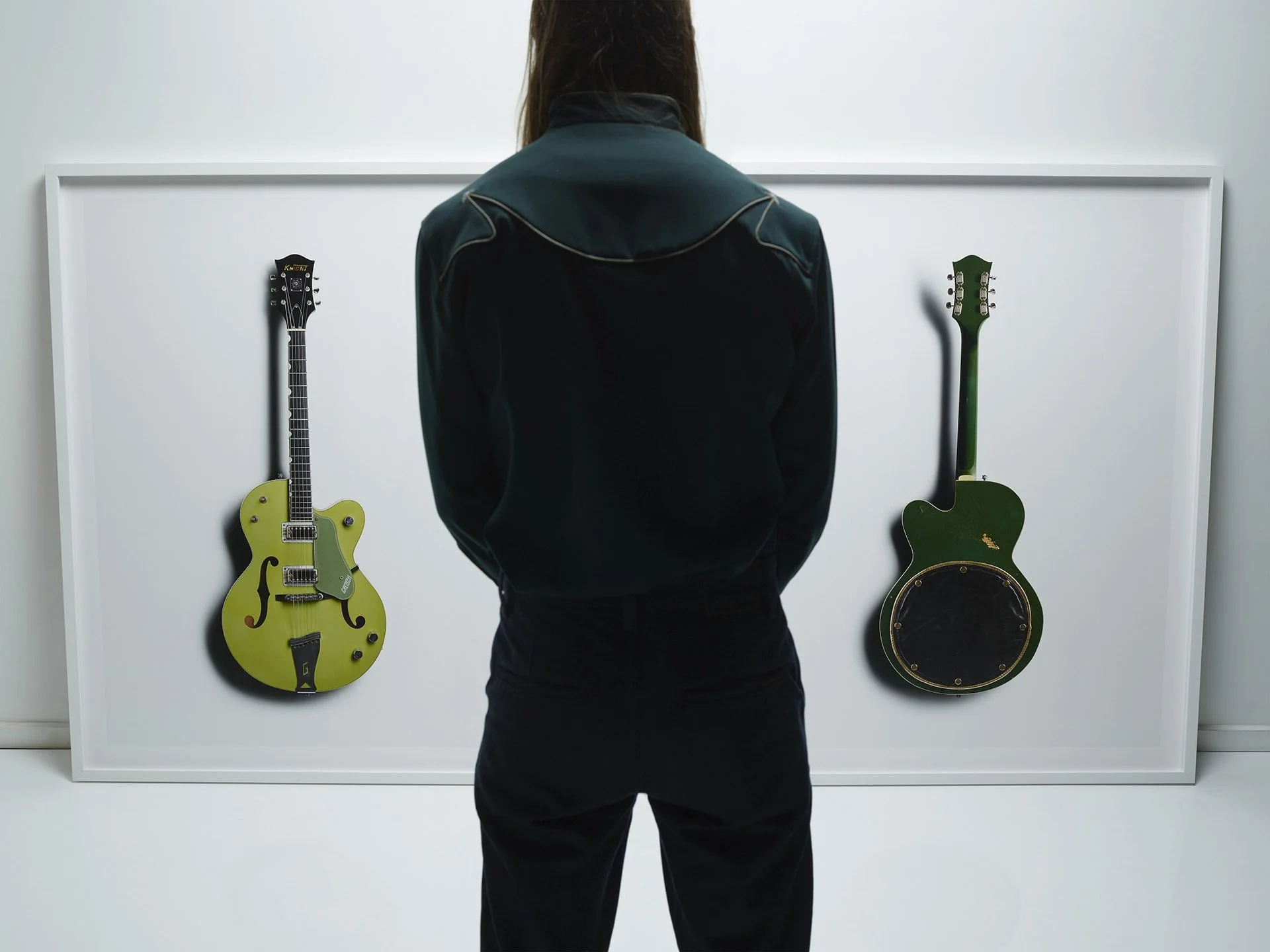 Person in black jacket looking at two guitars mounted on a white wall, one yellow with black accents and one green with black accents.