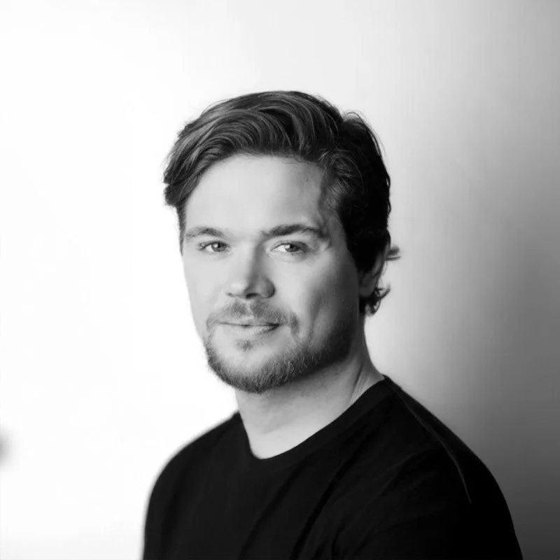 Black and white portrait of a young man with wavy hair, slight beard, and a confident expression, wearing a dark shirt, against a light background.