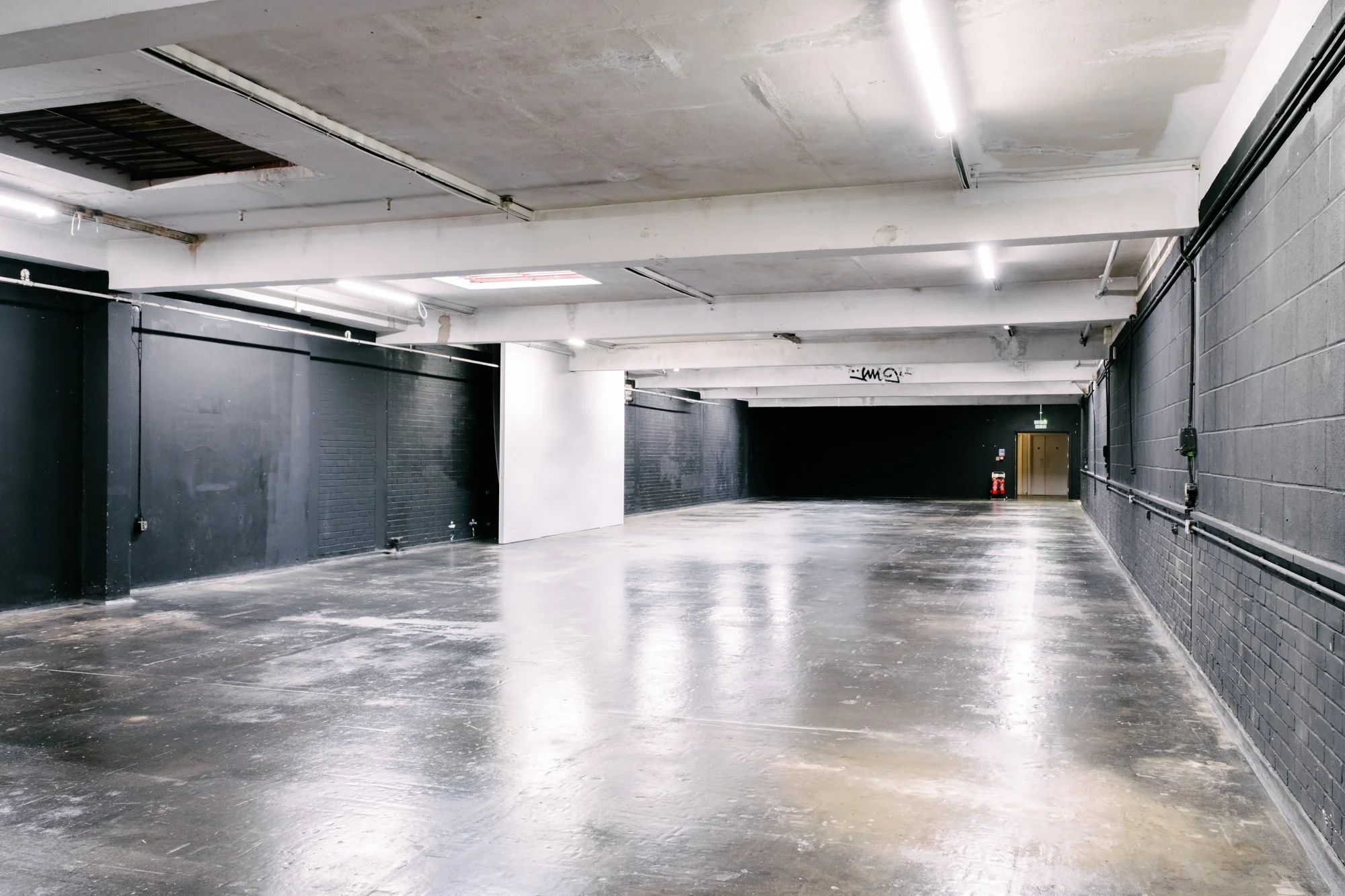 An empty indoor parking garage with black brick walls, dark polished concrete floors, and exposed piping on the walls. Fluorescent lights illuminate the ceiling, and a fire extinguisher is visible near a doorway at the back.
