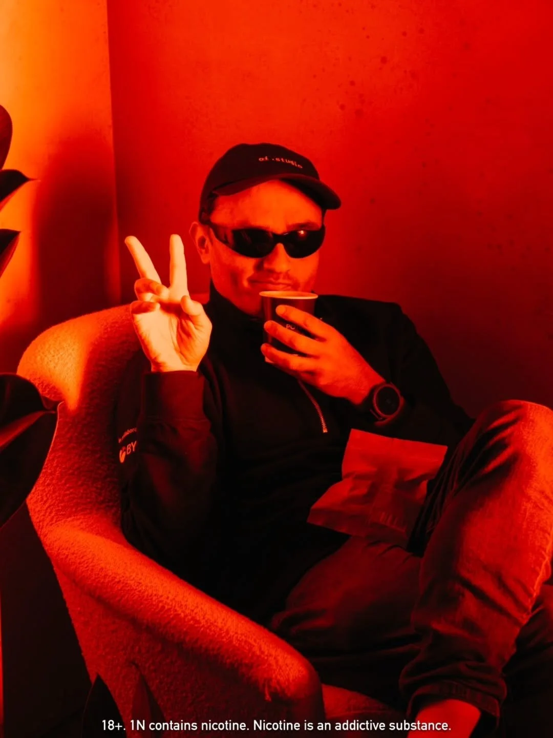 A man sitting in a chair, wearing sunglasses and a cap, holding a cup and making a peace sign with his hand.