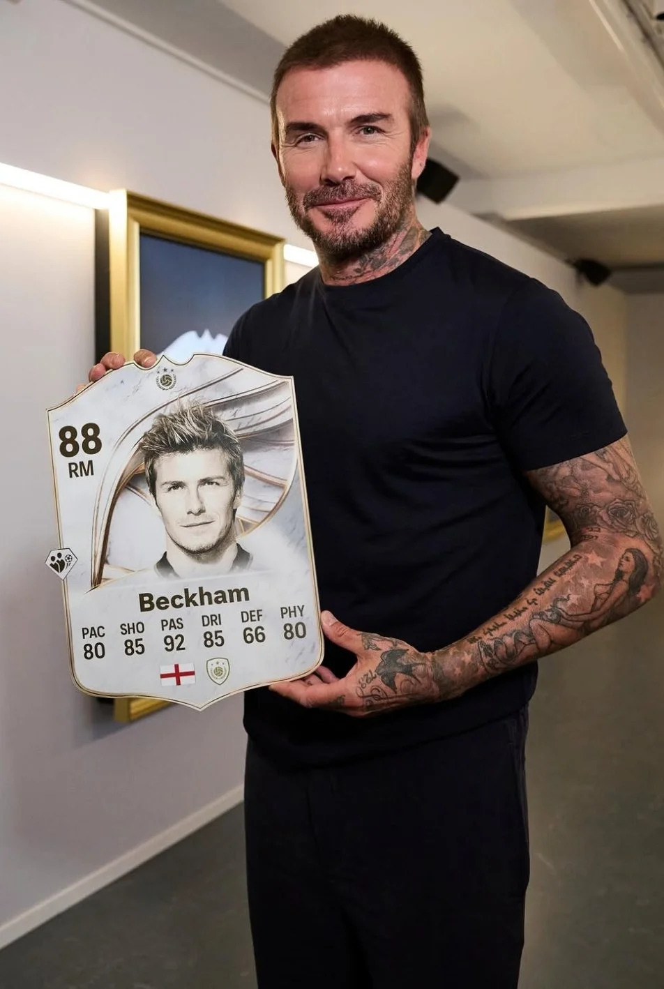 A man with tattoos on both arms and a beard holding a large FIFA-style card of David Beckham, featuring his portrait and soccer stats, in an indoor setting.
