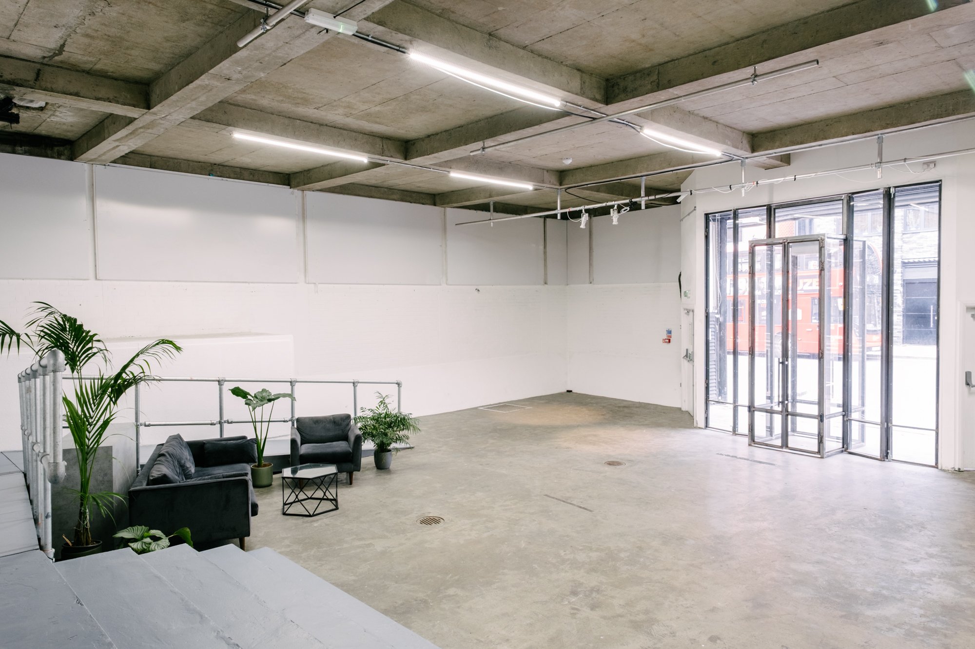 Interior of a modern, industrial-style space with a concrete ceiling and floor, white walls, large glass door, and minimal furniture including black sofas and potted plants.