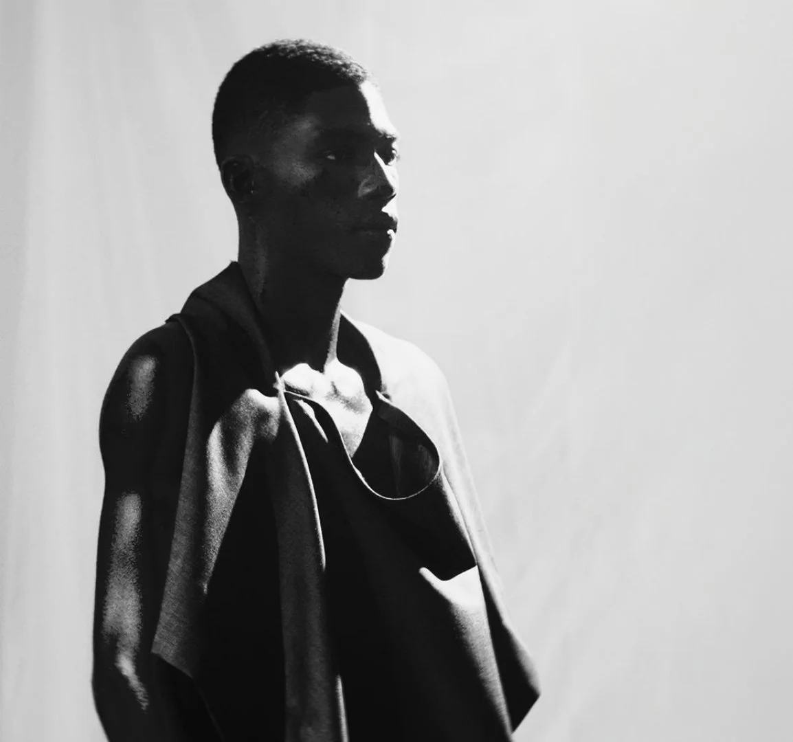 Black and white photo of a young person with short hair and a hoodie, standing against a plain light-colored background.