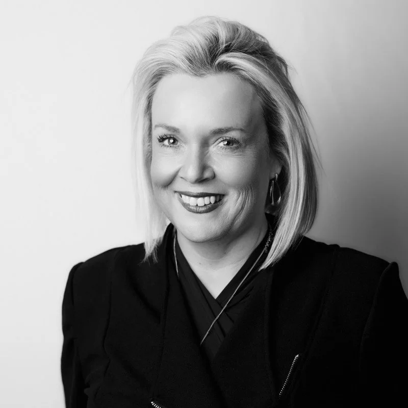 Black and white portrait of a smiling woman with shoulder-length blonde hair, wearing earrings and a dark outfit.
