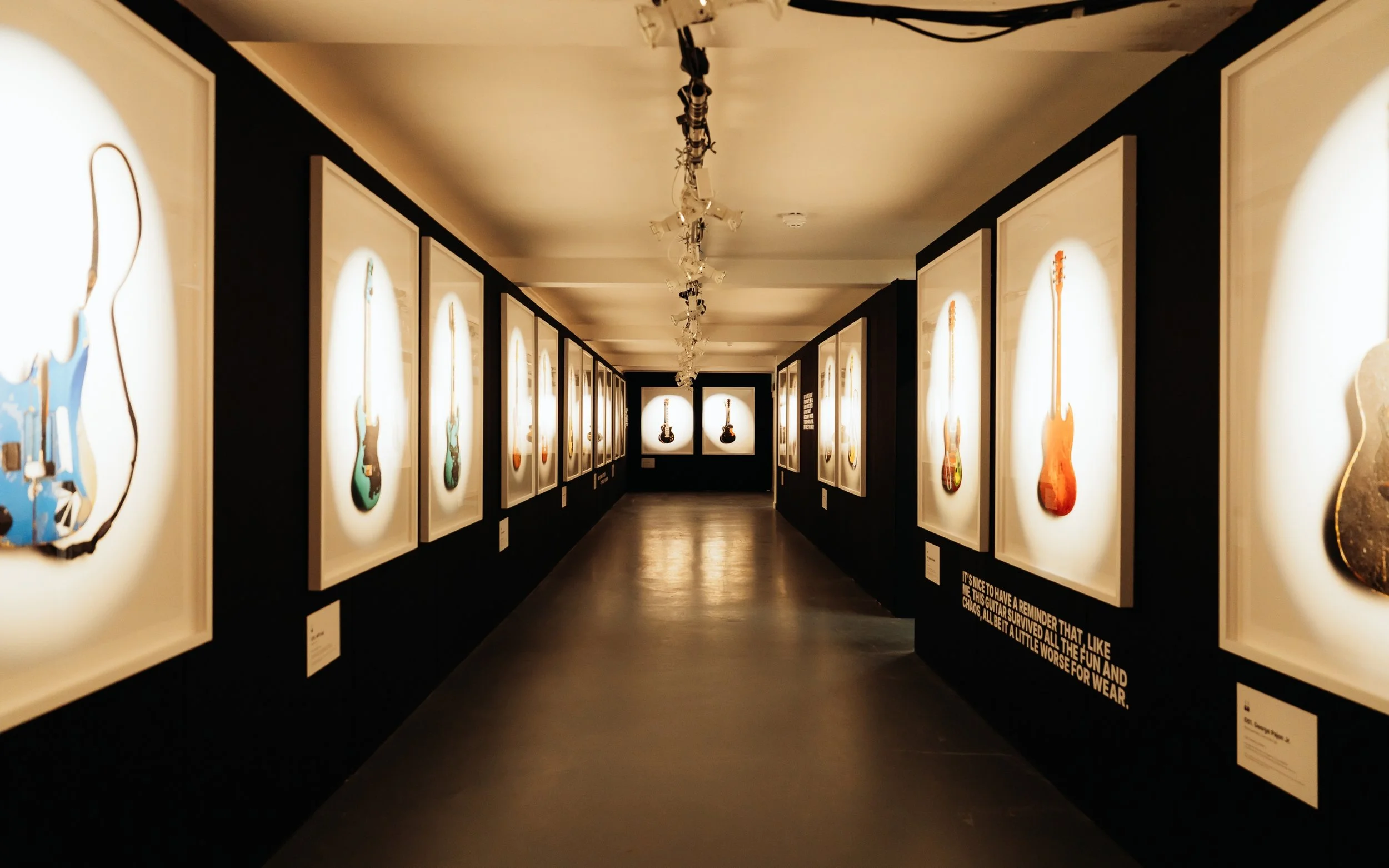 An art gallery with framed pictures of colorful guitars on black walls, illuminated by overhead gallery lighting.