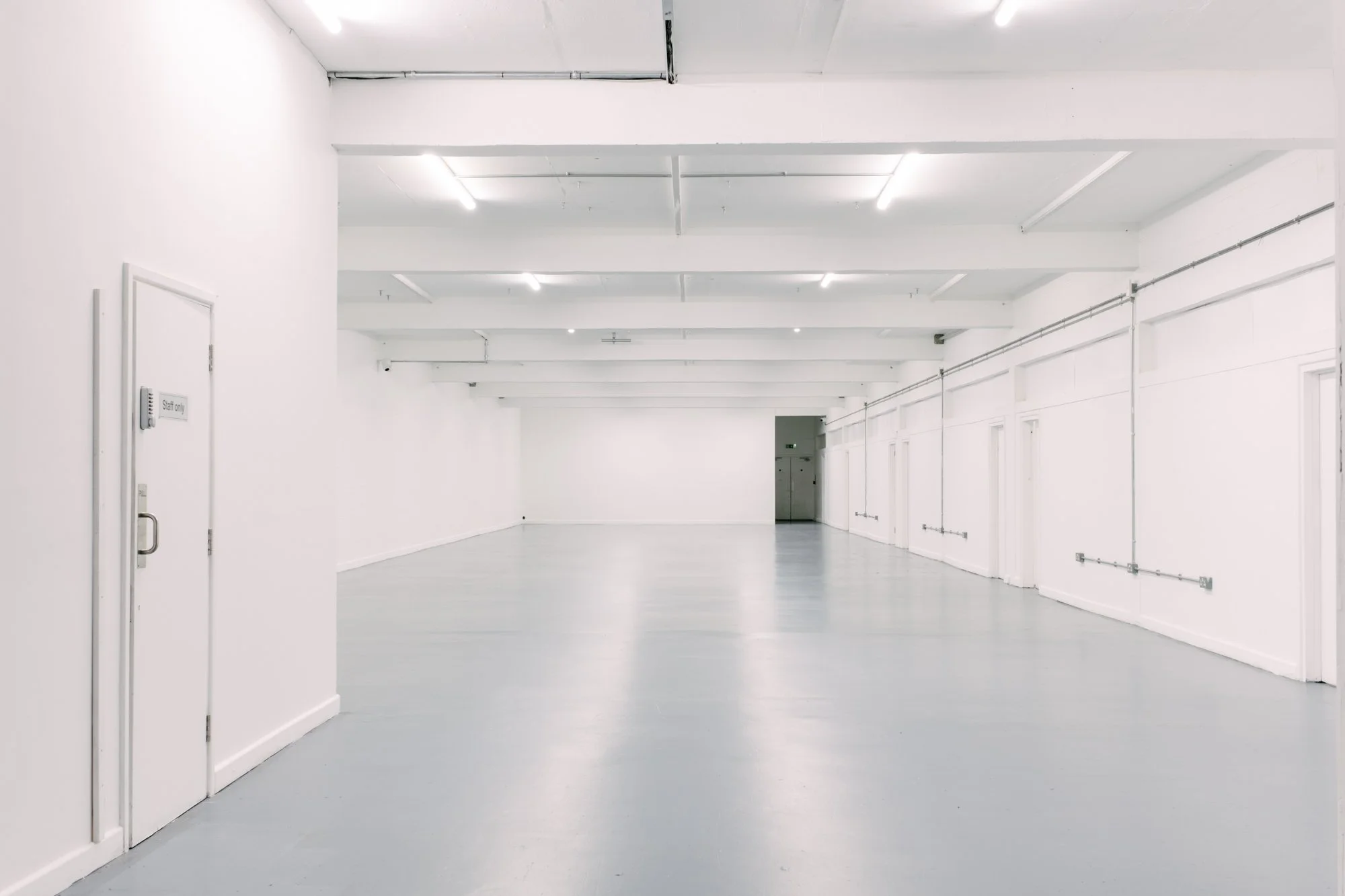 Empty white-walled corridor in a modern, minimalist building with fluorescent lighting and closed doors along the side.