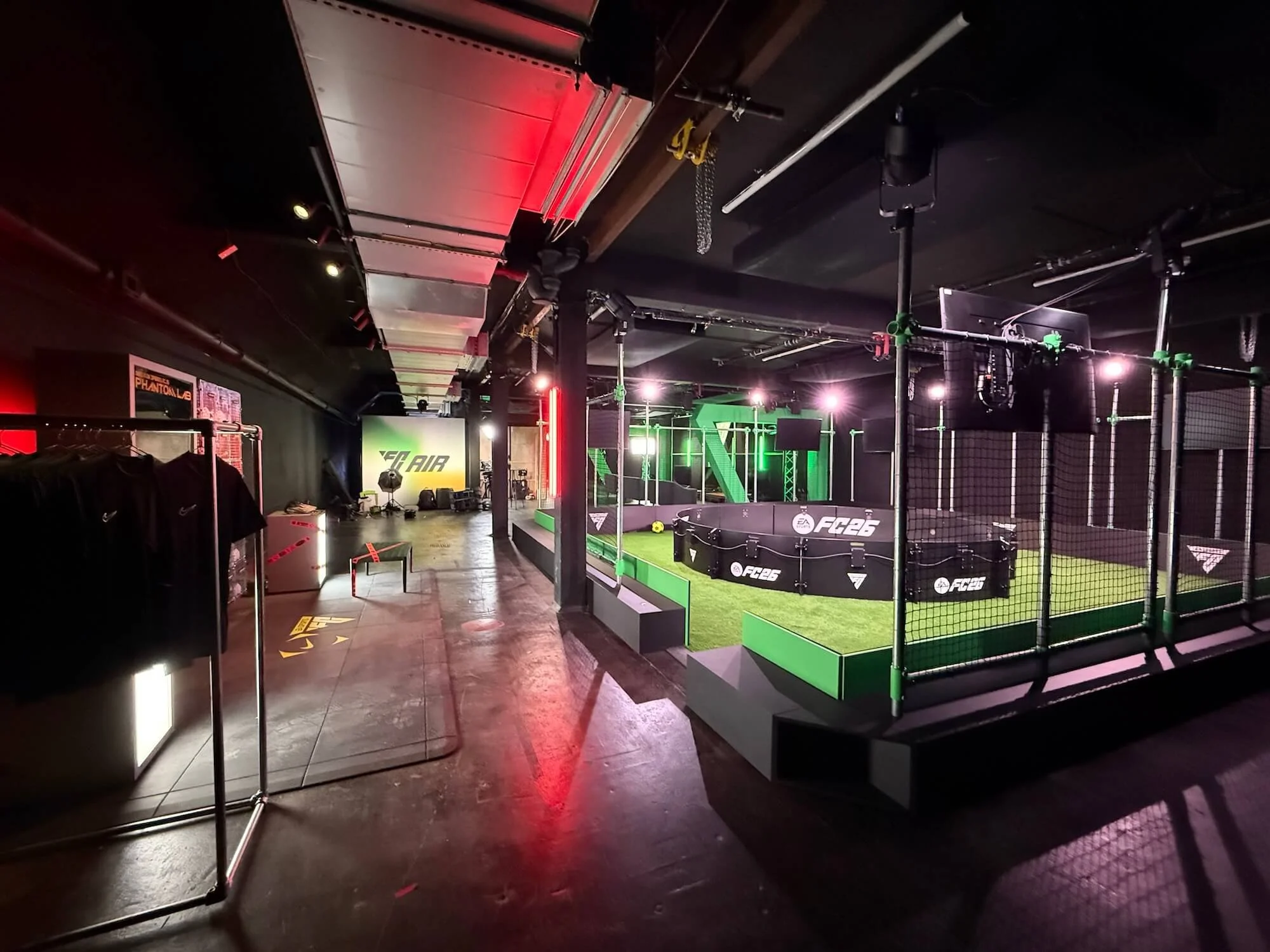 An indoor virtual reality game space with a soccer-themed setup, with a small soccer goal, bright lighting, and gaming equipment like monitors and barriers.
