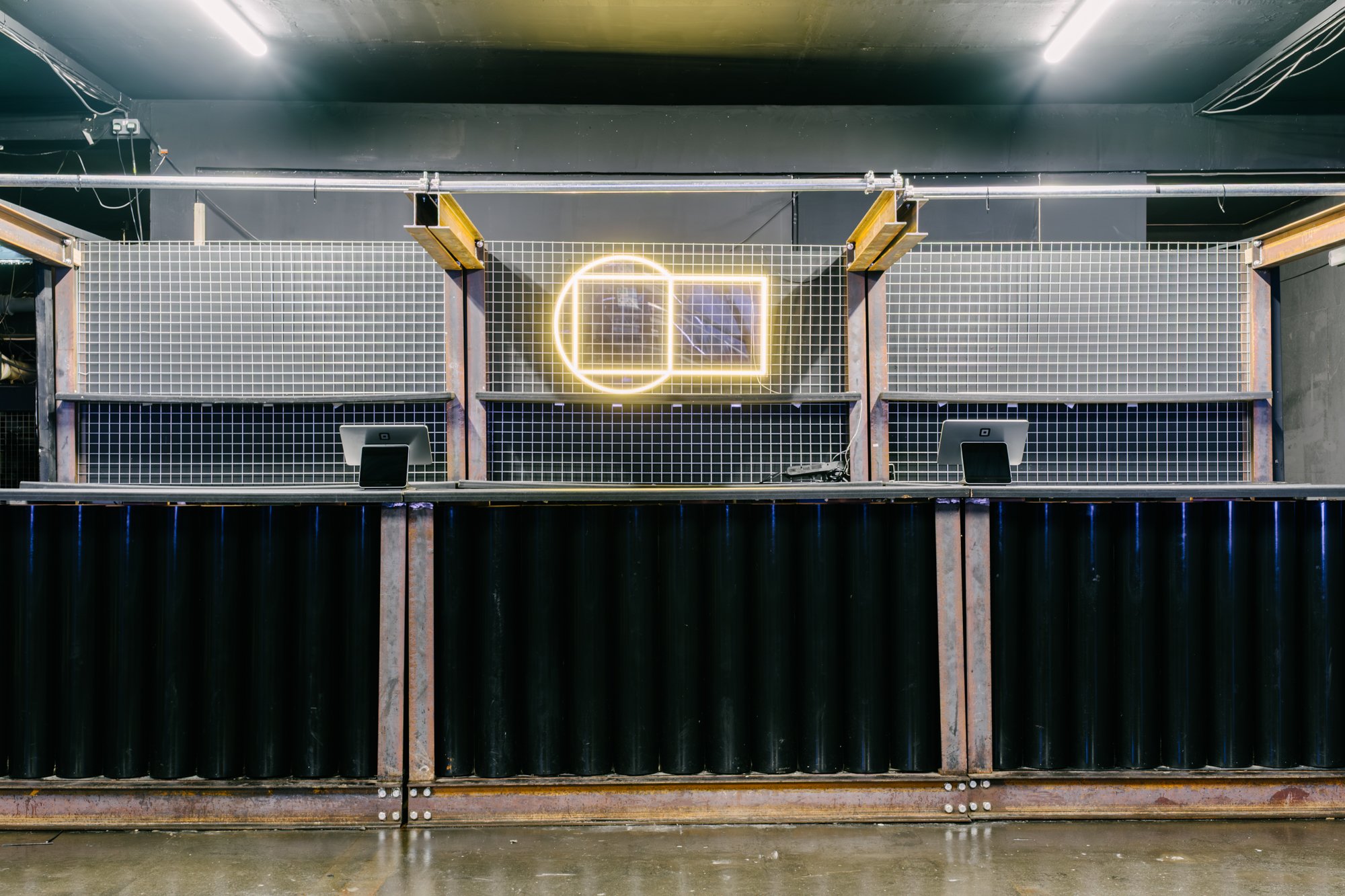 Interior view of an industrial space with a black ceiling, metal infrastructure, and a wall with a neon sign depicting a square and a circle with the square overlapping the circle. The space includes computer monitors and wires, and a corrugated metal fence in the foreground.