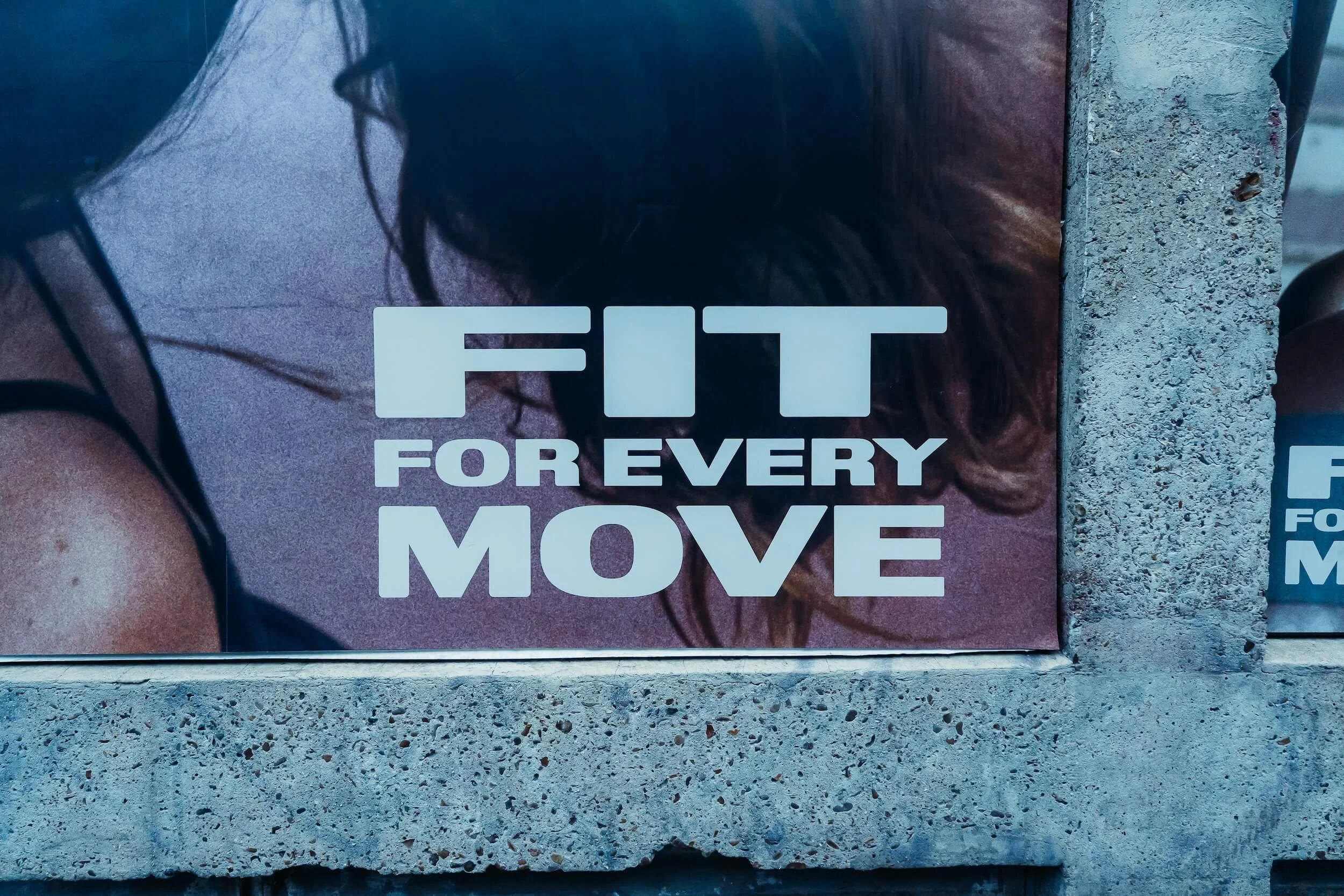 A poster on a concrete wall with the text 'FIT FOR EVERY MOVE' and a partially visible woman with dark hair and glasses.