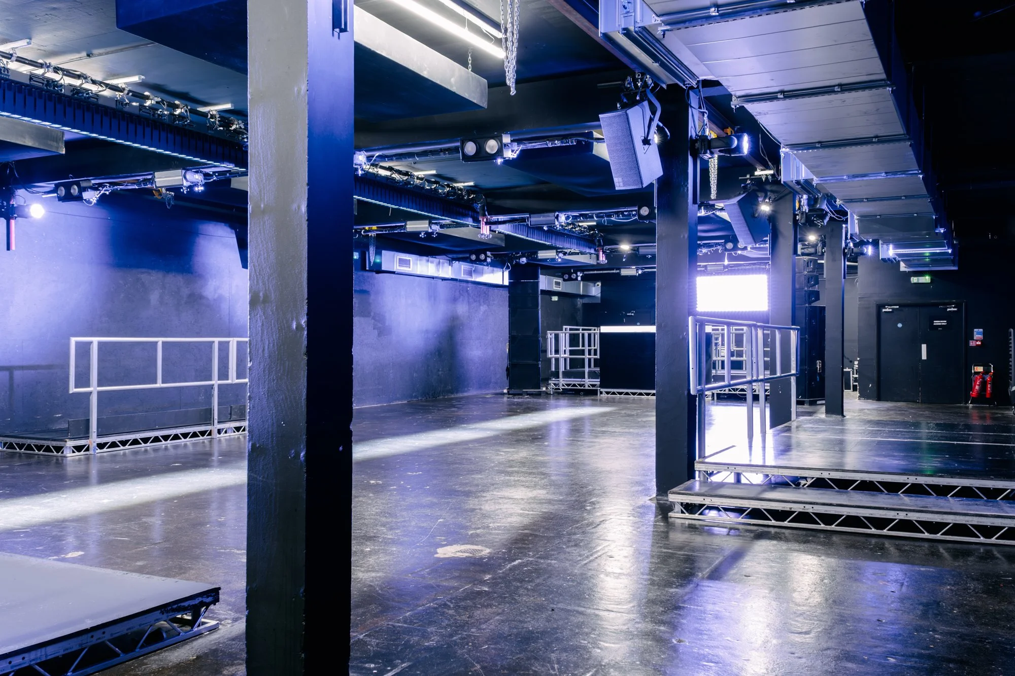 Empty stage or performance area with black walls, blue lighting, and metal scaffolding and equipment, possibly for a concert or event.