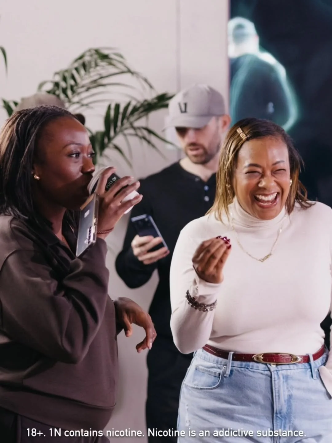 Three people are in a social setting, two women and one man. The woman on the left is drinking from a disposable coffee cup, while the woman on the right is laughing with her eyes closed. The man in the background is looking at his phone. The scene appears lively and casual, with a plant and a dark abstract art piece in the background.