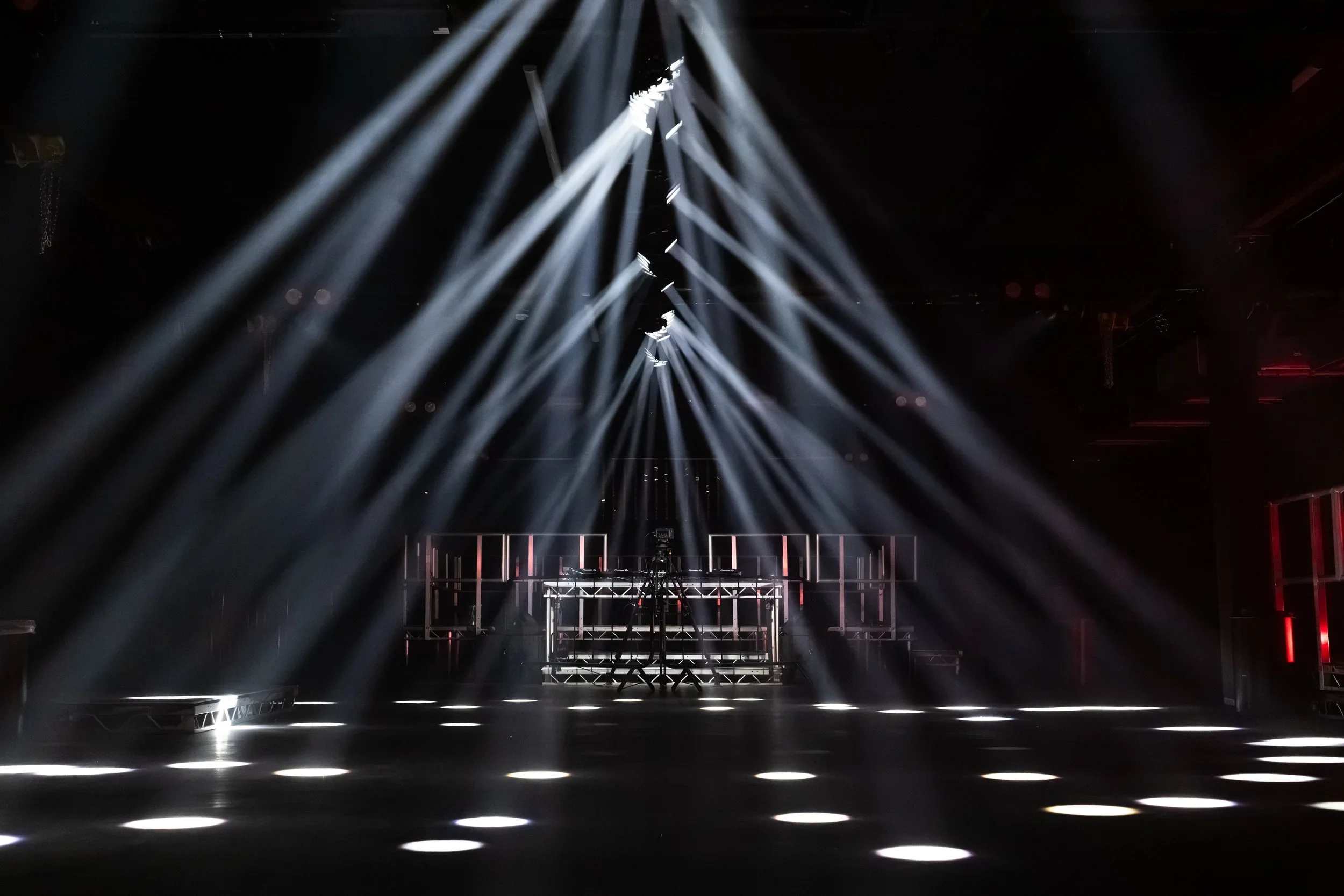 Dark stage with multiple bright white spotlights overhead, illuminating the floor with circular light patterns, and a DJ booth or performance area in the background.