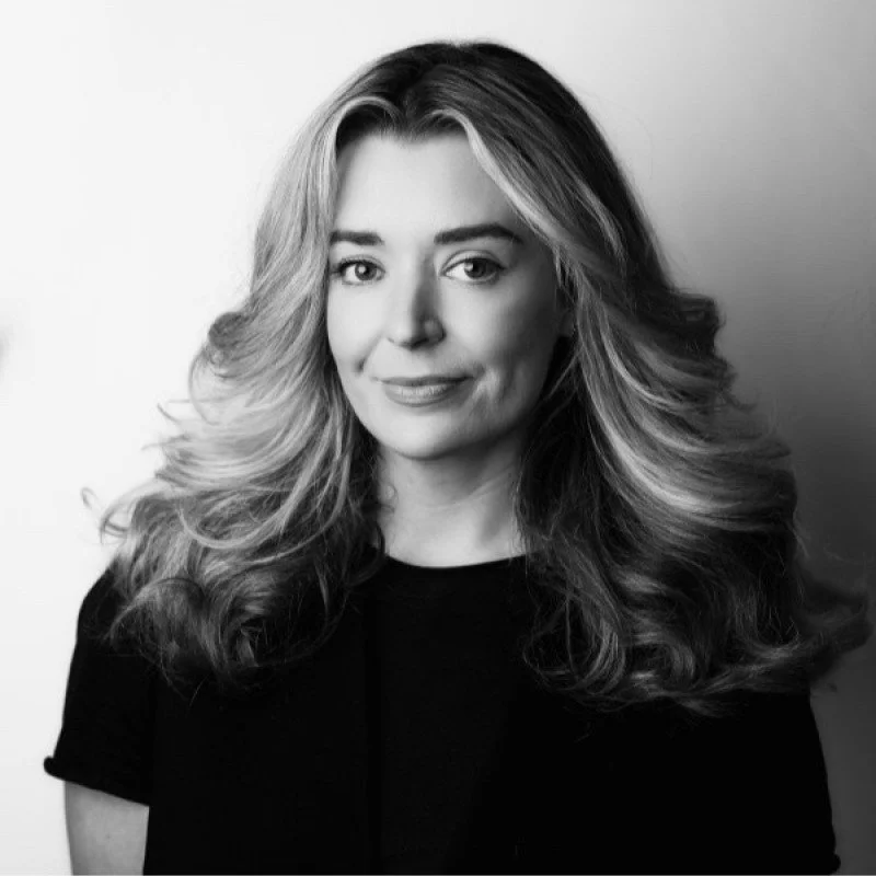 Black and white portrait of a woman with wavy, shoulder-length hair, wearing a black top, looking at the camera with a slight smile.