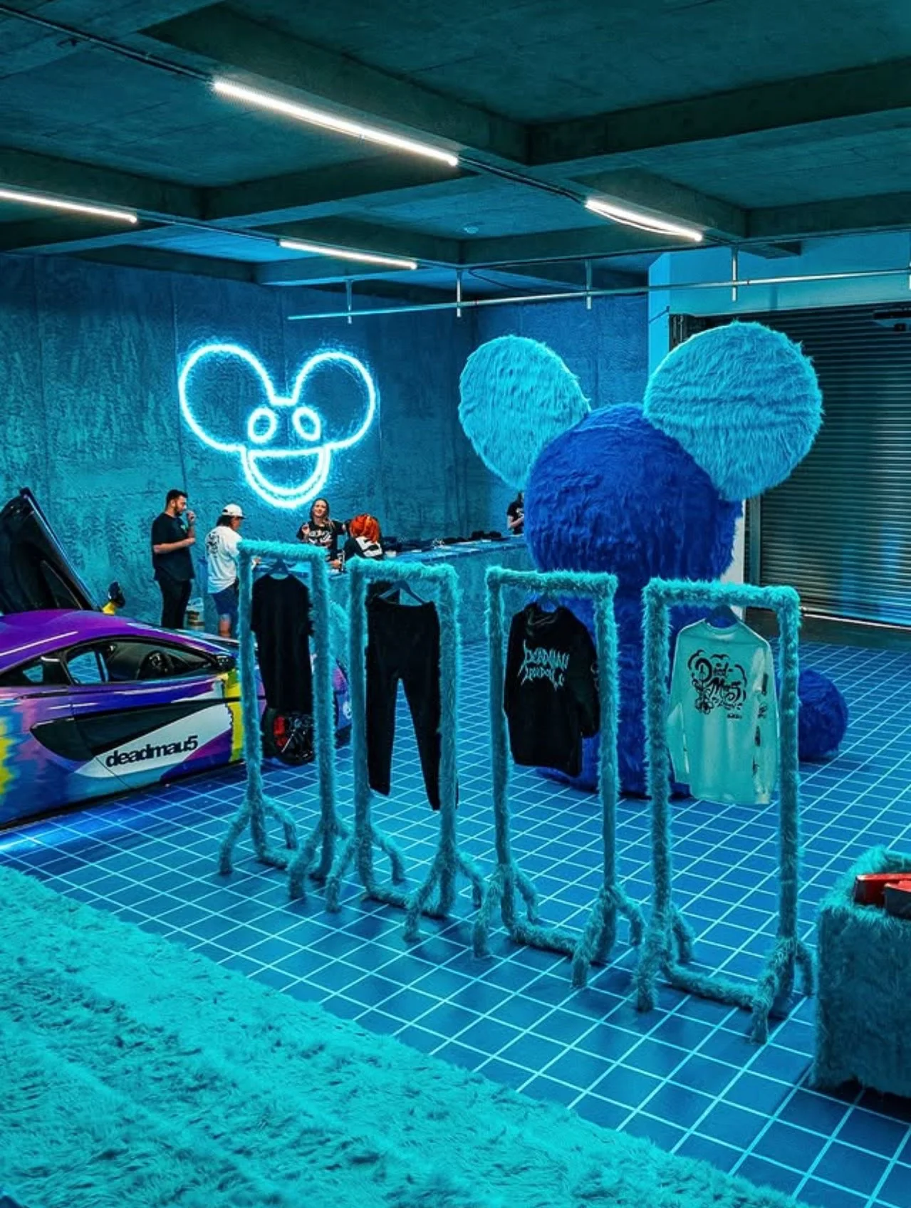 An indoor space with a neon Mickey Mouse head outline on the wall, a large fuzzy blue Mickey Mouse head sculpture, clothing racks with apparel, and a purple race car.