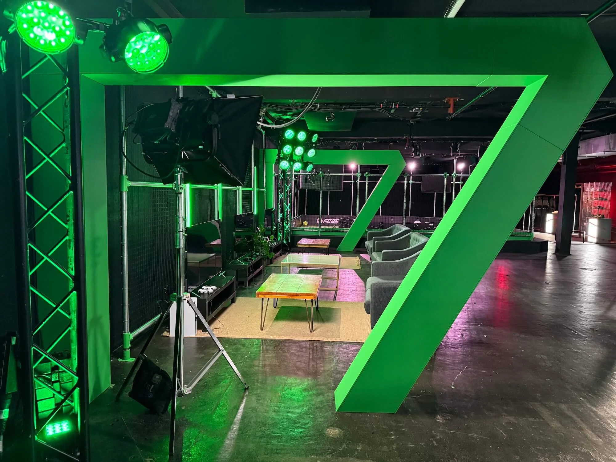 An indoor sports facility with green lighting and large green geometric structures resembling the number seven. There are chairs, tables, and a netted area for sports activities.