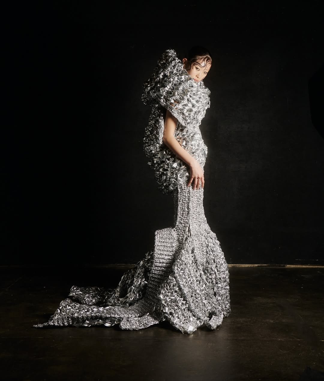 Model wearing an avant-garde silver metallic gown with textured embellishments, standing against a dark background.