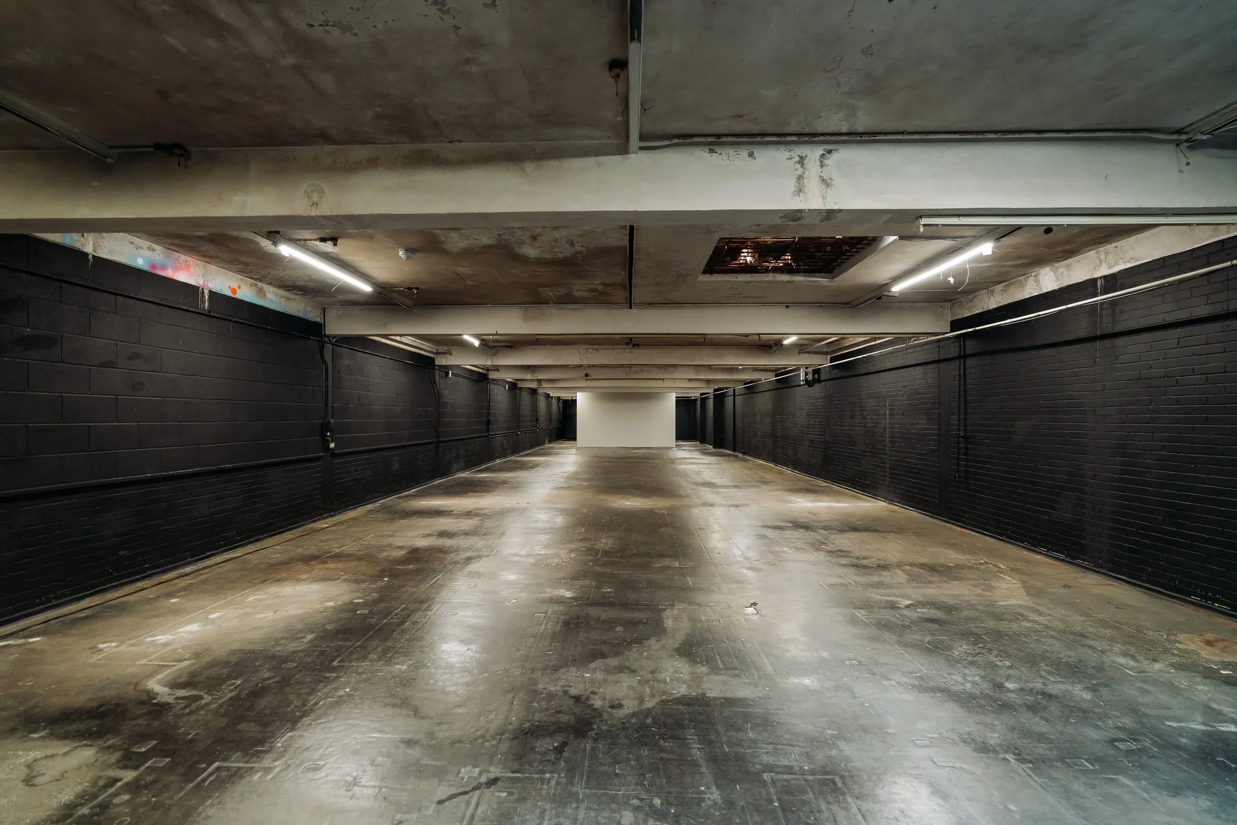 Empty underground parking garage with black walls, concrete ceiling, and a white wall at the end.