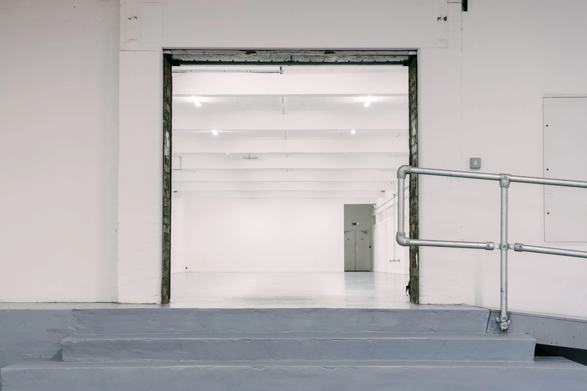 An indoor space with white brick walls, a large open doorway, and gray steps leading up to an open area. The space features exposed pipes and minimal decor, giving it an industrial or modern look.