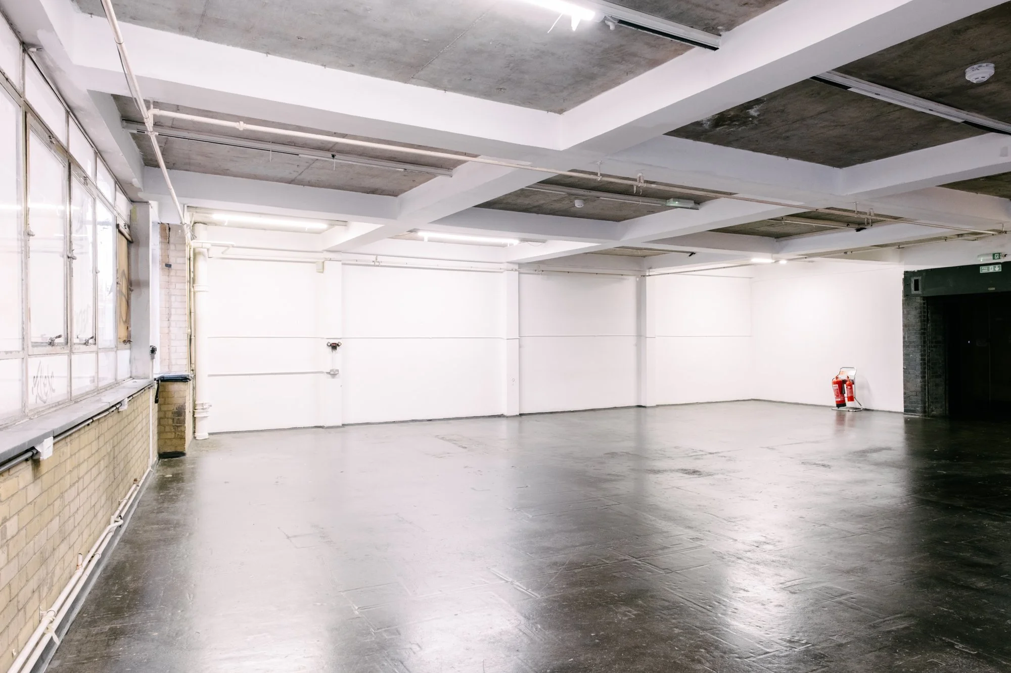 Empty indoor space with concrete floor, white and gray walls, visible piping, and a fire extinguisher in the corner.
