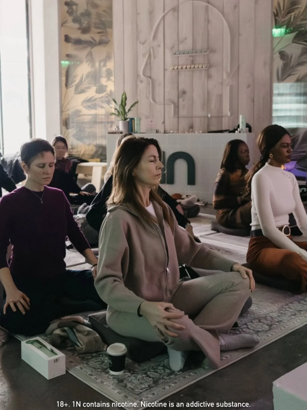 Group of women meditating indoors on a carpeted floor with eyes closed in a yoga class, with a wooden background and plants.