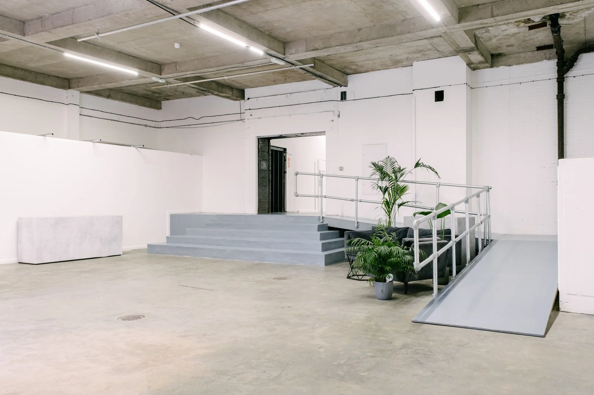 Interior of a modern, minimalist building with exposed concrete ceiling, white walls, and large open space. There are steps leading up to a doorway, a small plant near the stairs, and some furniture like a couch and white blocks.