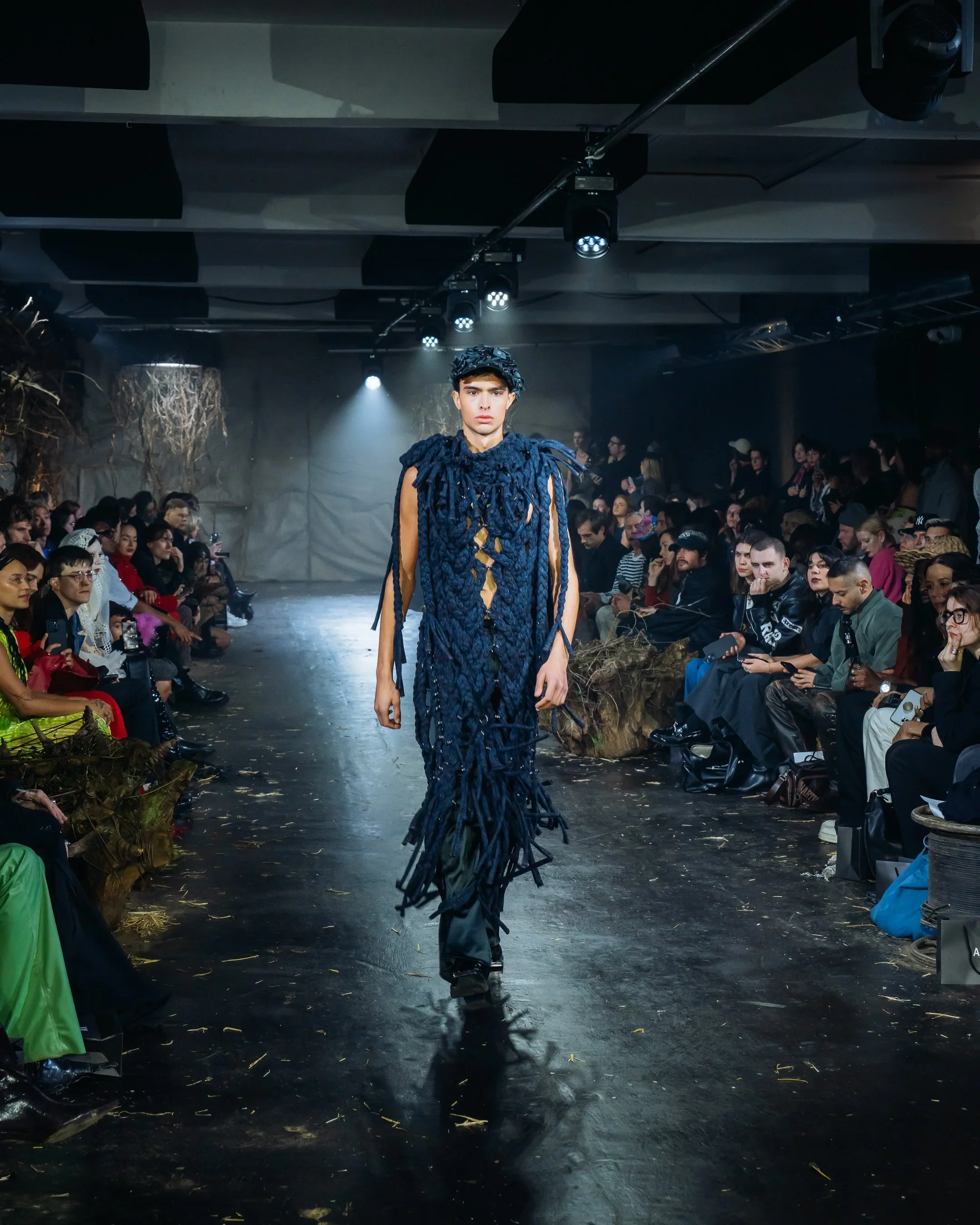 Model walking runway in dark blue, textured, dreadlock-inspired fashion design at a fashion show with audience seated on both sides.