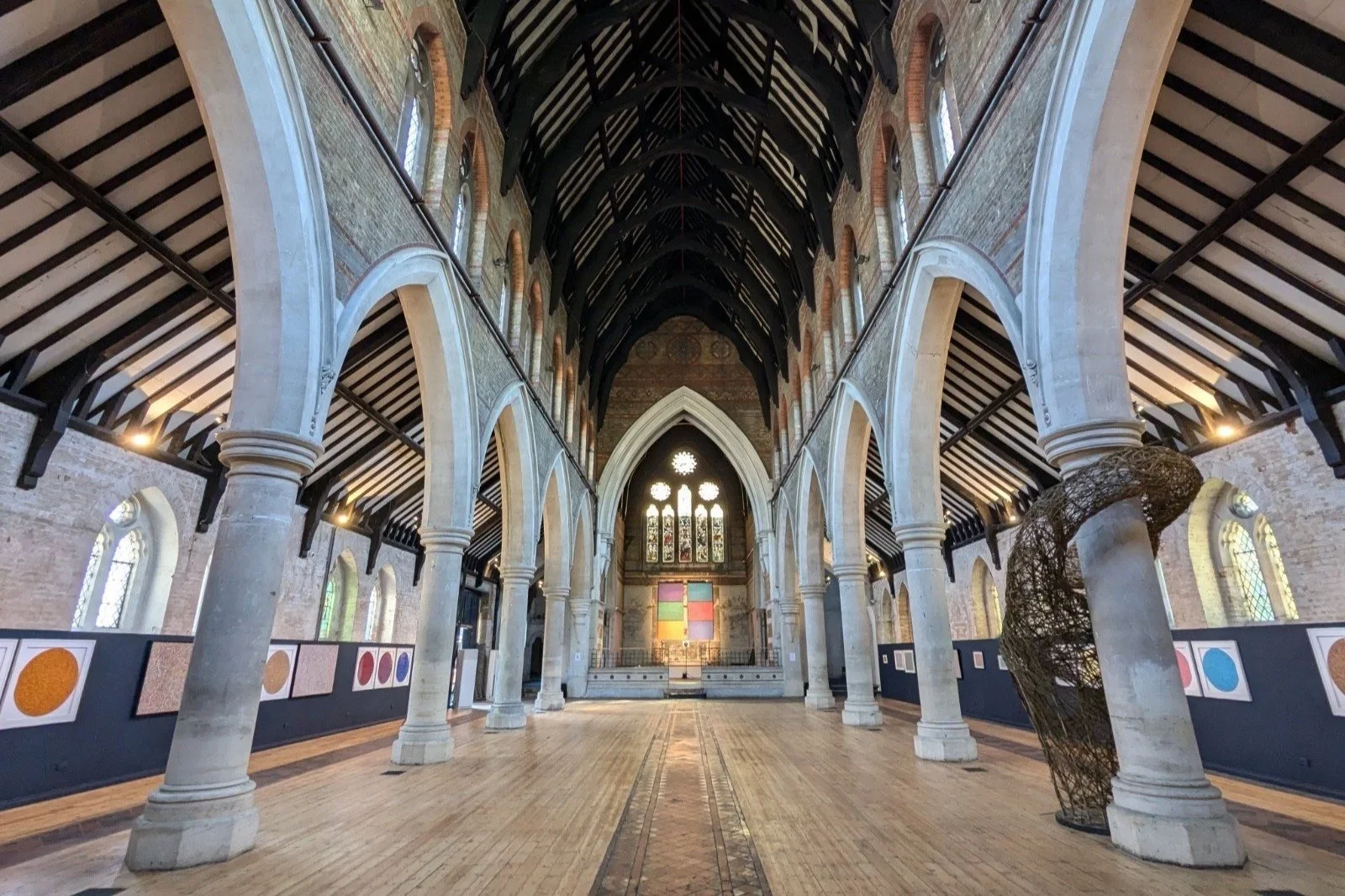 Interior of a church with high vaulted ceiling, stone arches, stained glass windows, wooden flooring, and an abstract sculpture near one pillar.