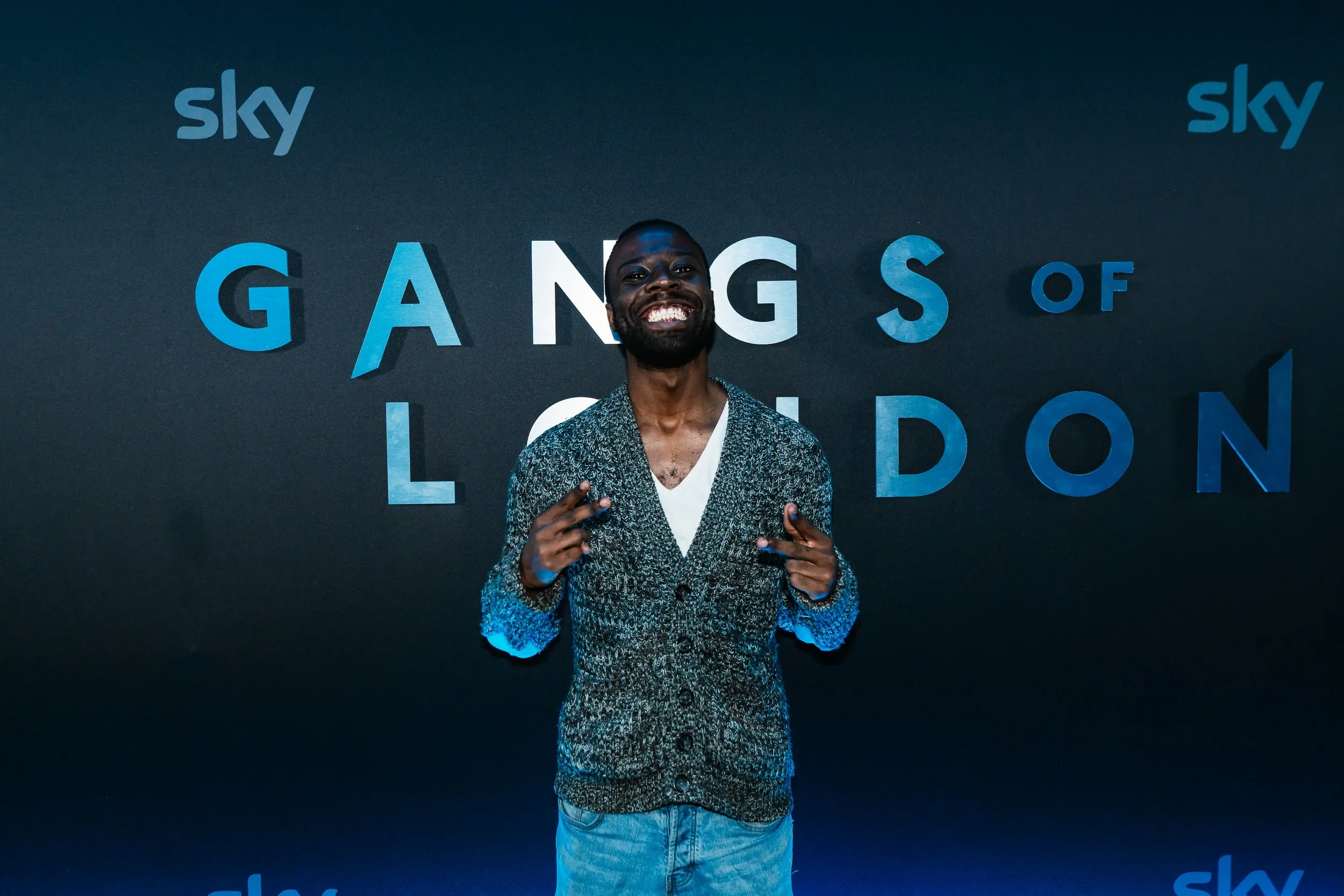 A man smiling and pointing with both hands in front of a black background with blue and white text that reads 'Gangs of London' and 'Sky' logo.