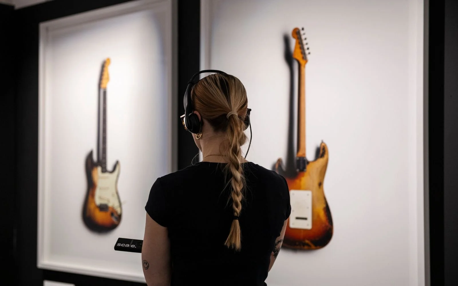 A woman with a braid and headphones looking at two electric guitars on display in a museum.