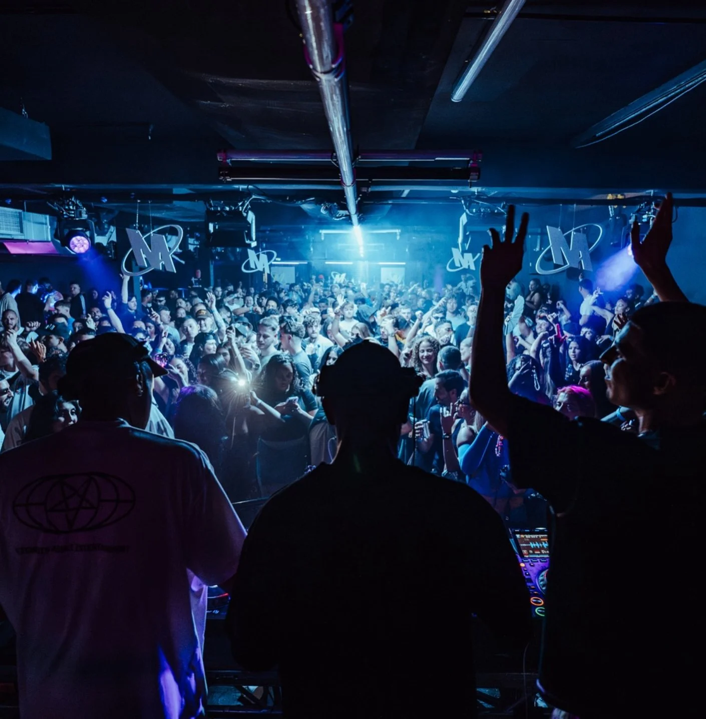 Crowded nightclub dance floor with people dancing and DJ in the foreground, blue lighting, and decorative elements such as hanging symbols and neon signs.