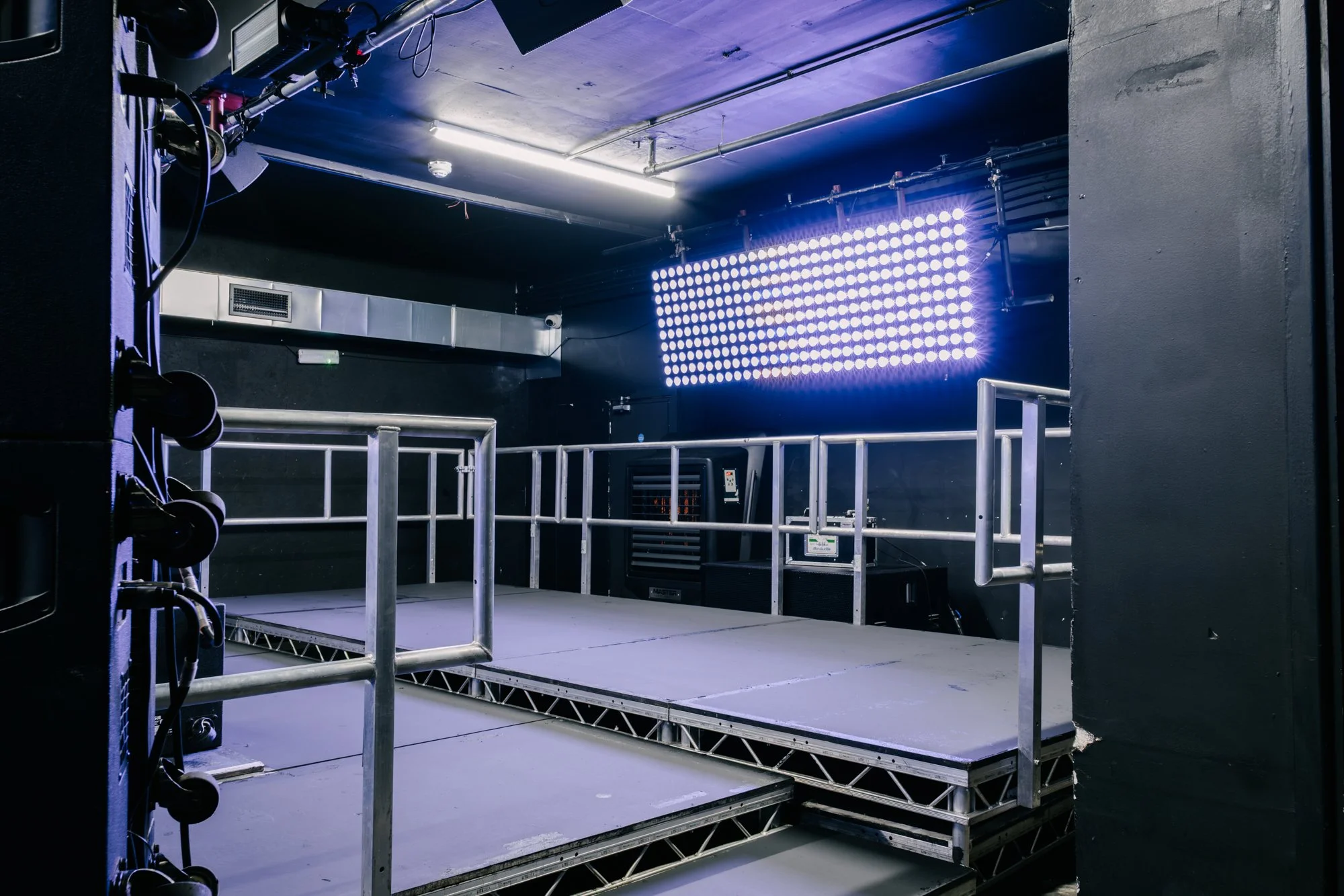 A stage with a metal railing and a large LED light panel on the wall, set up for a performance or event, in a dark room with industrial-style ceiling.