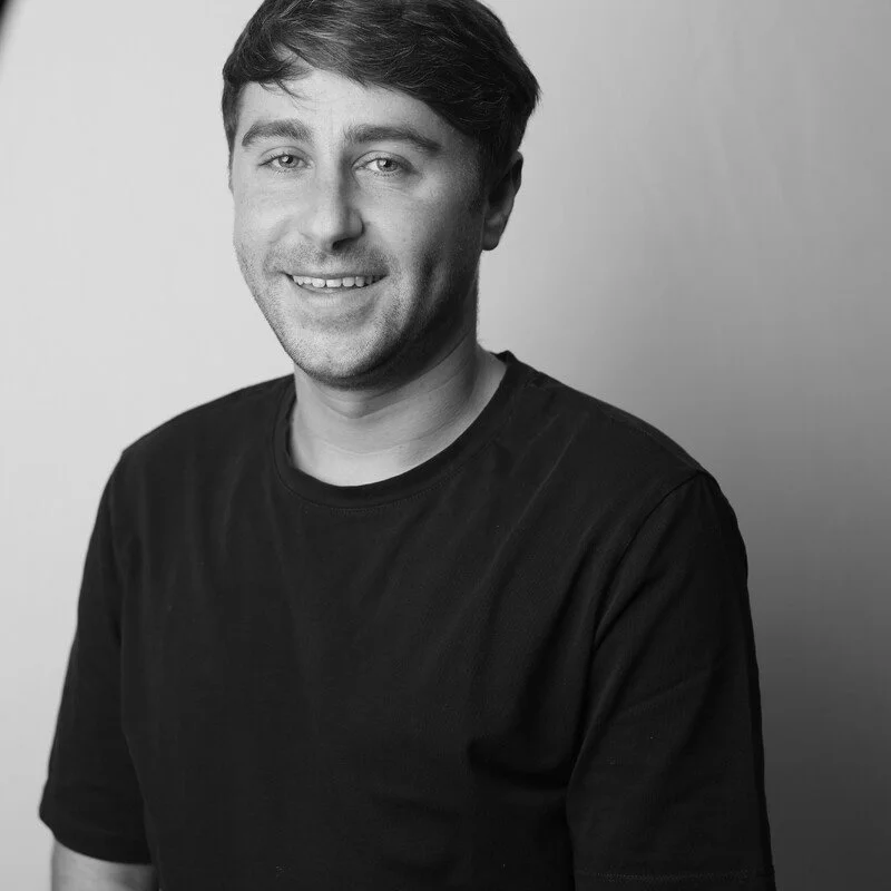 Black and white portrait of a smiling young man with short dark hair, wearing a plain black t-shirt, against a plain background.