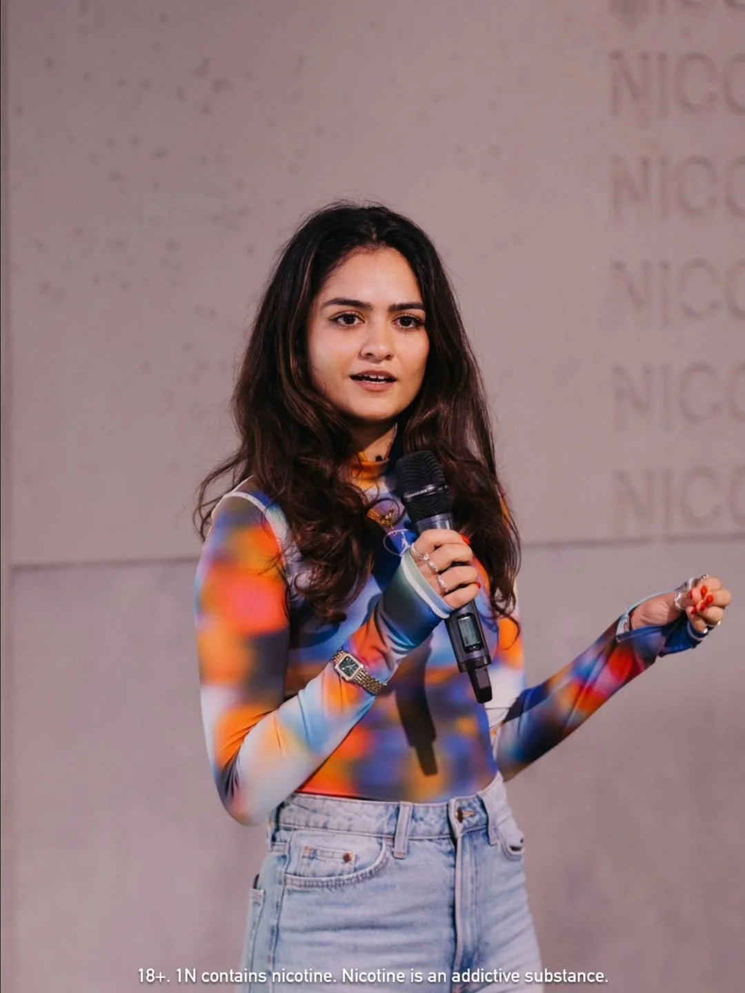 Young woman with long, wavy dark hair, wearing a colorful, long-sleeve blouse and light jeans, speaking into a microphone at an indoor event.