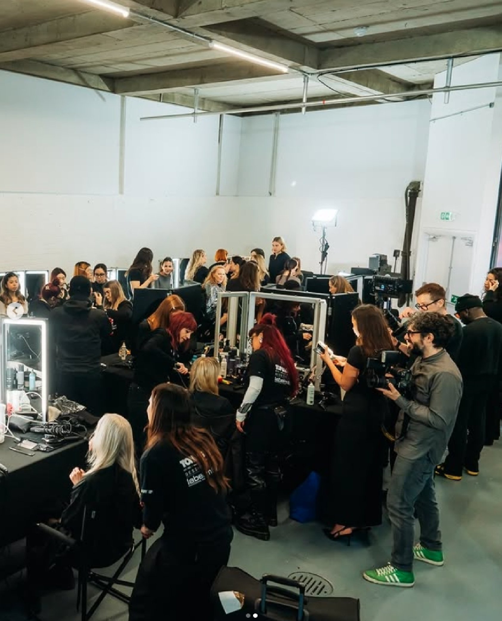 A crowded room with multiple tables and mirrors where many women are getting makeup done. Several people are taking photos or filming, and a photographer is holding a camera. The room has a high ceiling with exposed pipes and bright lighting.