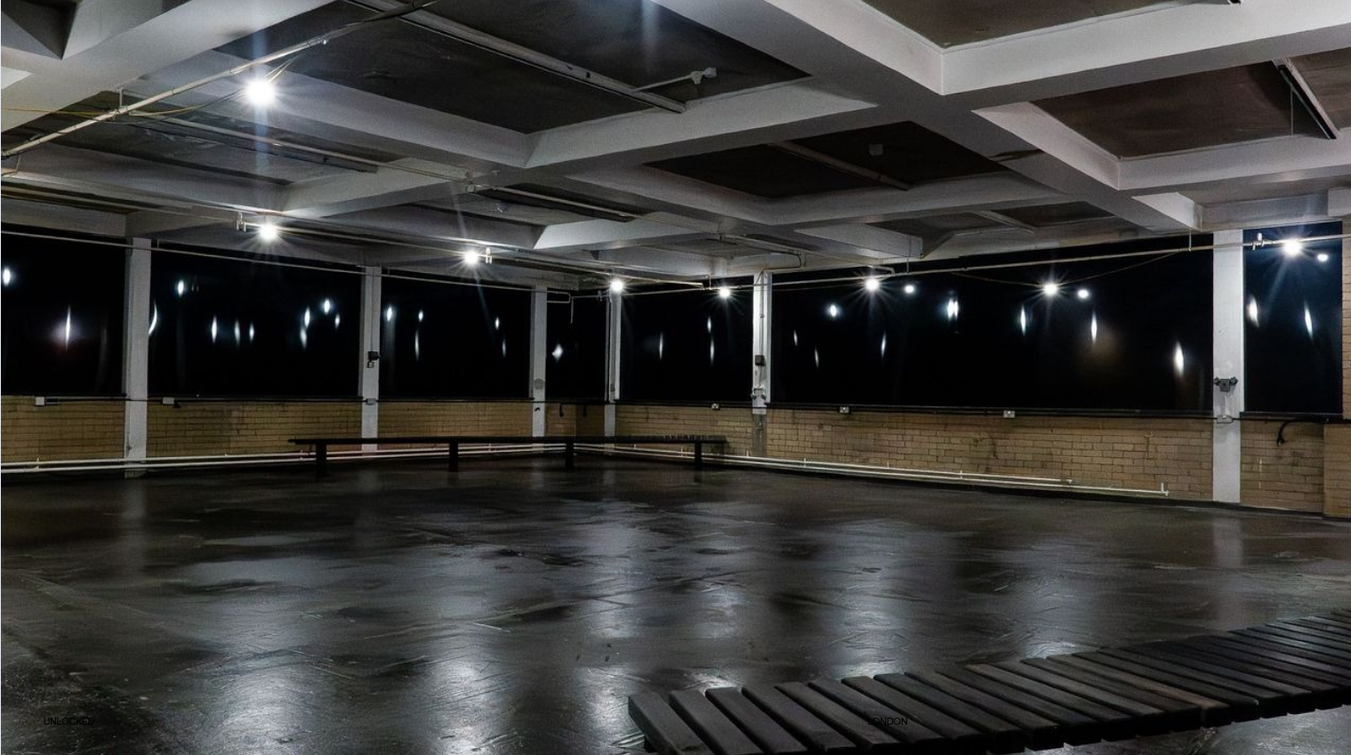Empty rooftop parking lot at night, illuminated by ceiling lights with a wet asphalt surface and brick walls.