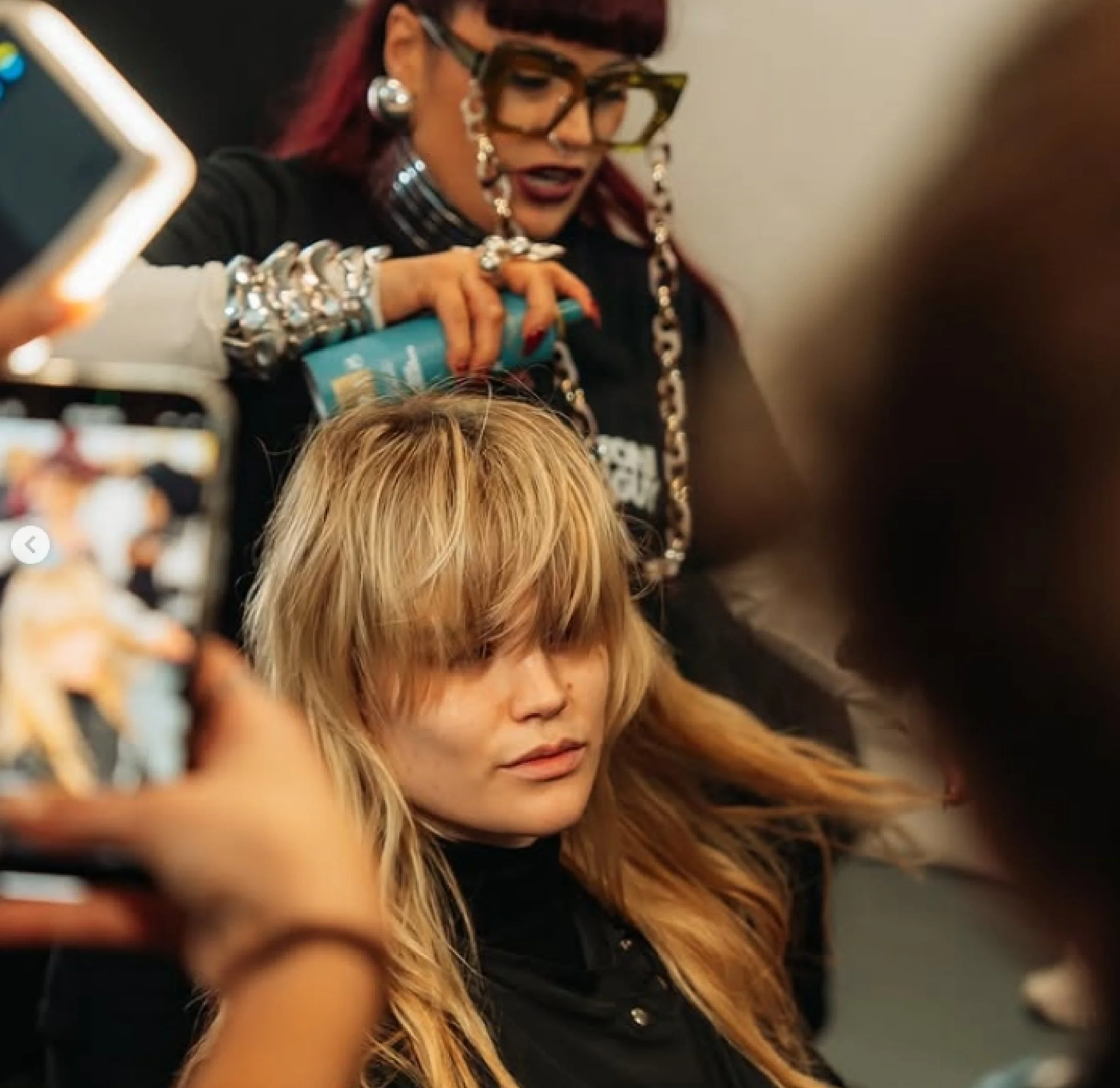 A woman with long blonde hair and bangs sits with eyes closed as she gets her hair styled, while another woman with large glasses, bright jewelry, and colorful hair spray styles her hair at a busy salon or backstage setting.