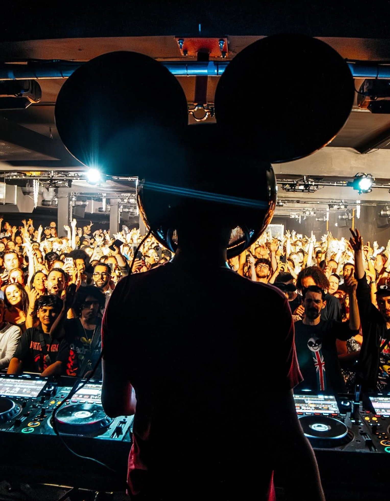 Silhouette of a DJ with Mickey Mouse ears helmet performing at a concert, with a crowd of people dancing and raising their hands in the background under stage lights.