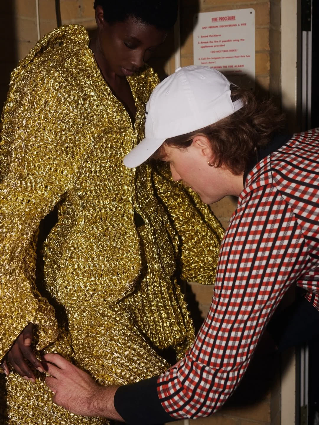 A person adjusting the hem of a golden, textured dress worn by a woman in a white cap and checkered shirt.