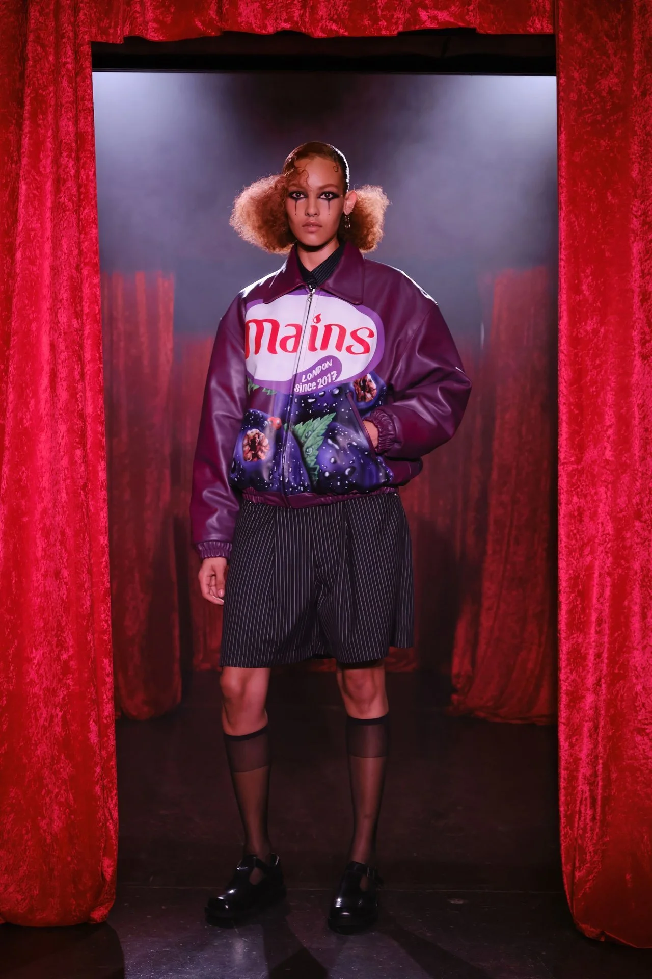 Model standing on stage with red velvet curtains, wearing a purple bomber jacket with space and nature graphics, black pinstripe shorts, sheer black socks, and black shoes, with dramatic makeup and curly hair.