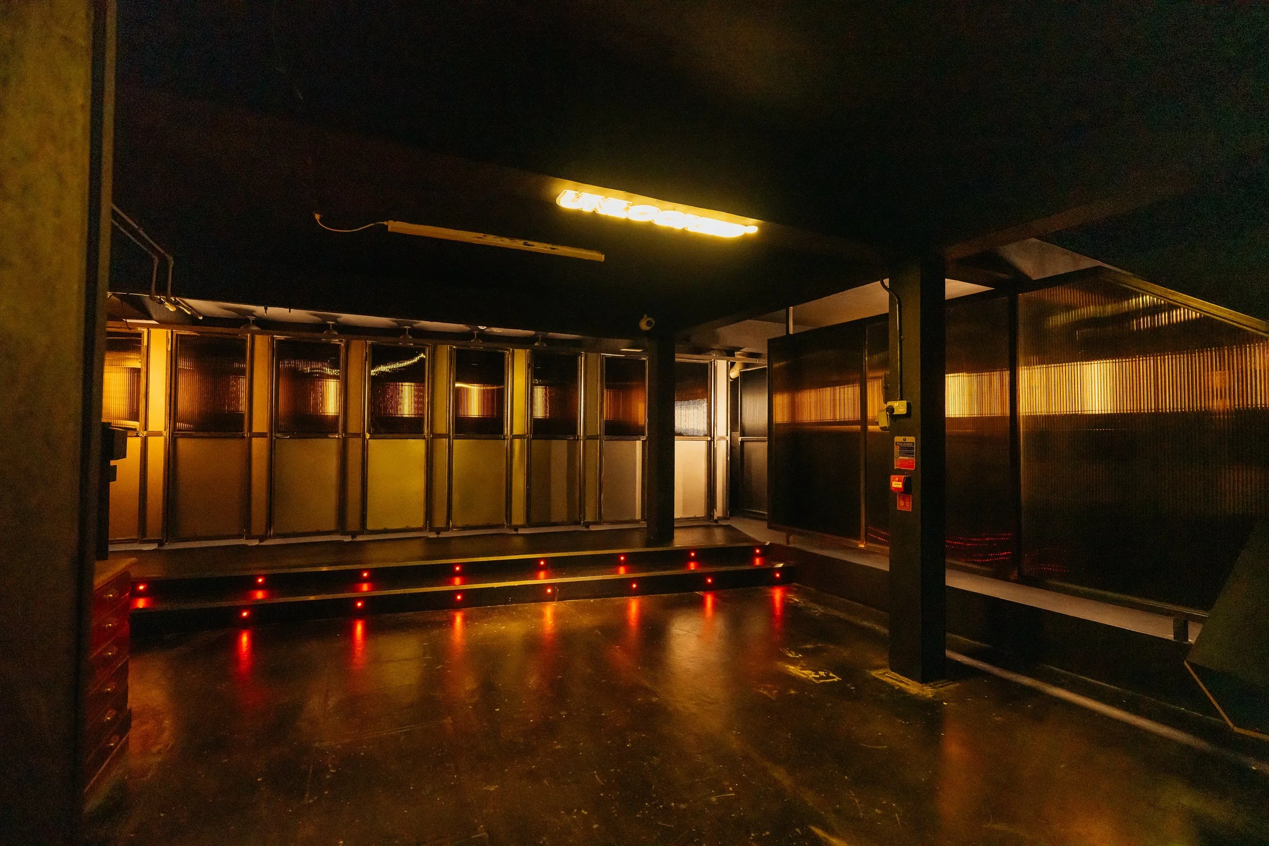 Empty nightclub dance floor with red LED lights along the bottom, a black ceiling with a yellow neon sign, and a back wall with decorative panels illuminated with golden light.