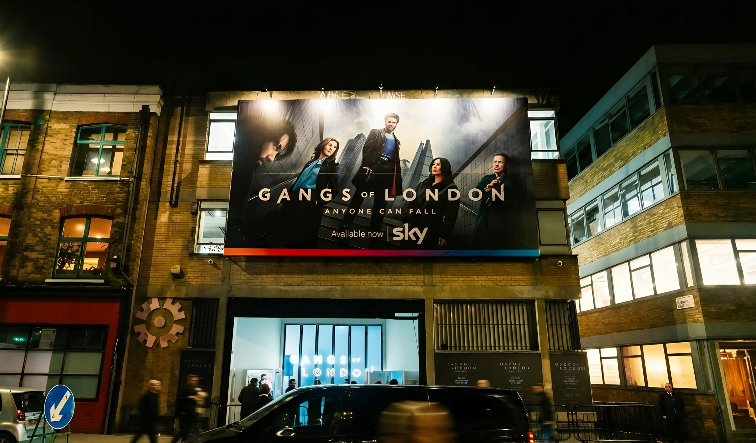 Nighttime city street with a large illuminated billboard advertising a TV show called 'Gangs of London' on Sky, featuring five characters and the tagline 'Anyone Can Fall.'