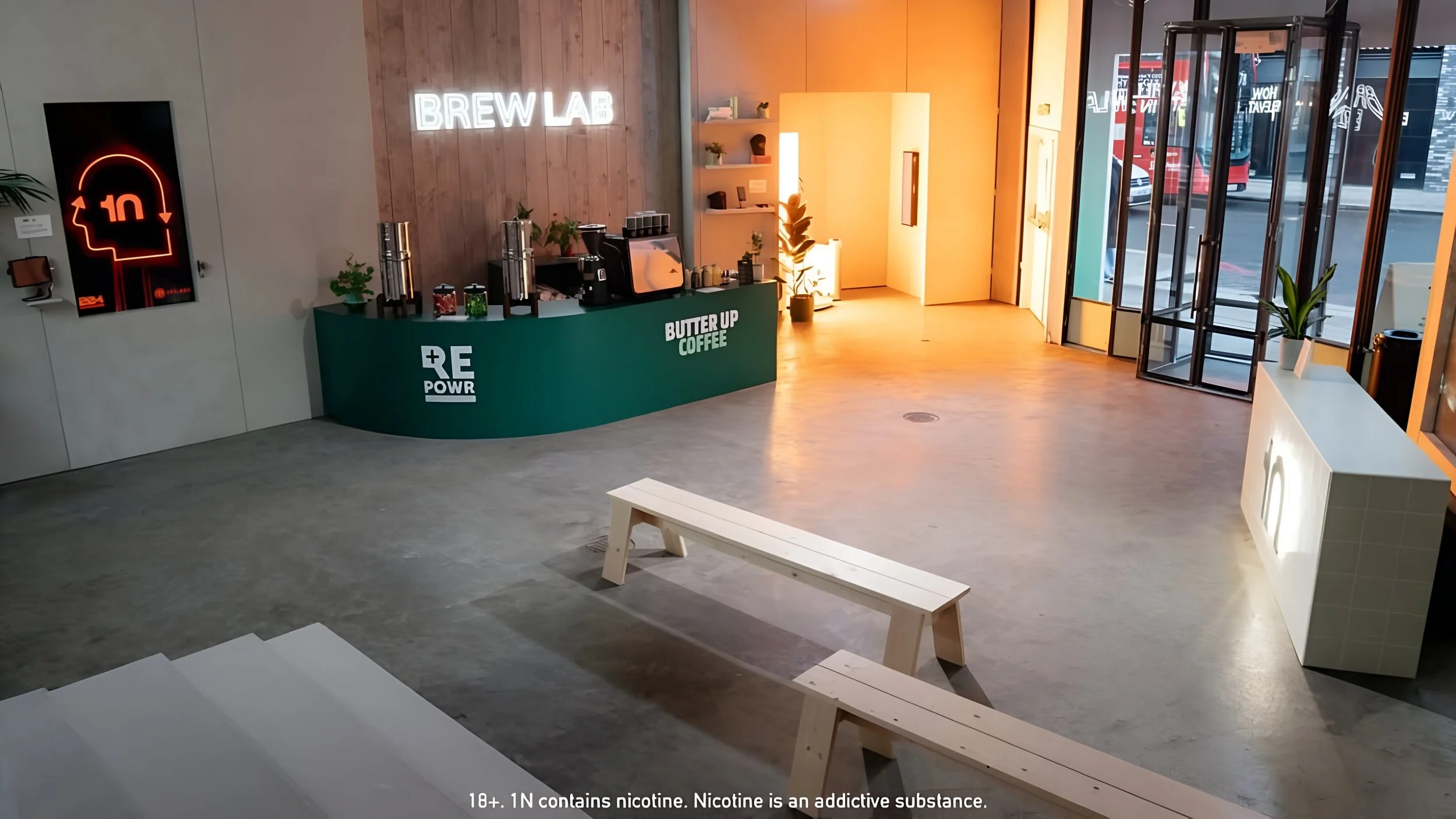 Interior of a modern coffee shop with a green counter labeled 'Butter Up Coffee' and 'RE POWR'. There are coffee machines and supplies on the counter, a neon sign on the wooden wall that says 'BREW LAB', and a digital sign with a red outline of a per