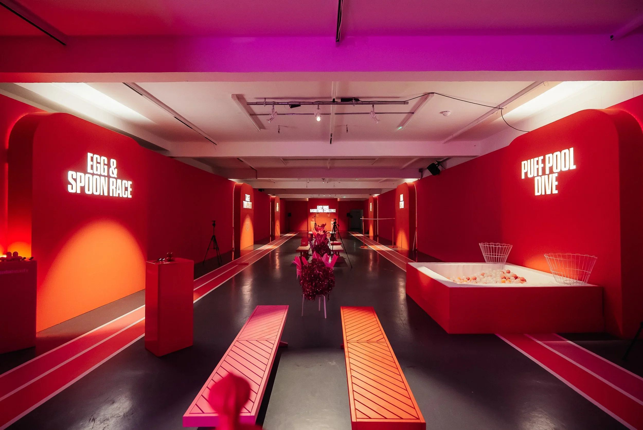 An indoor room with red walls and dim lighting. There are signs on the walls indicating different activities such as 'Egg & Spoon Race' and 'Puff Pool Dive.' The floor has designated lanes with white lines, and there are benches, tripods, and a ball 