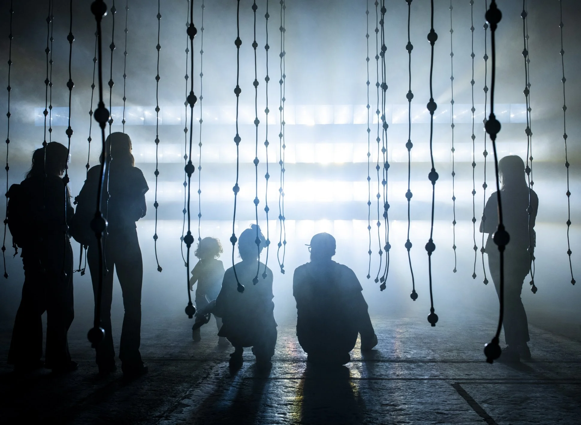 People silhouette behind hanging black bead strings with bright foggy light in the background.