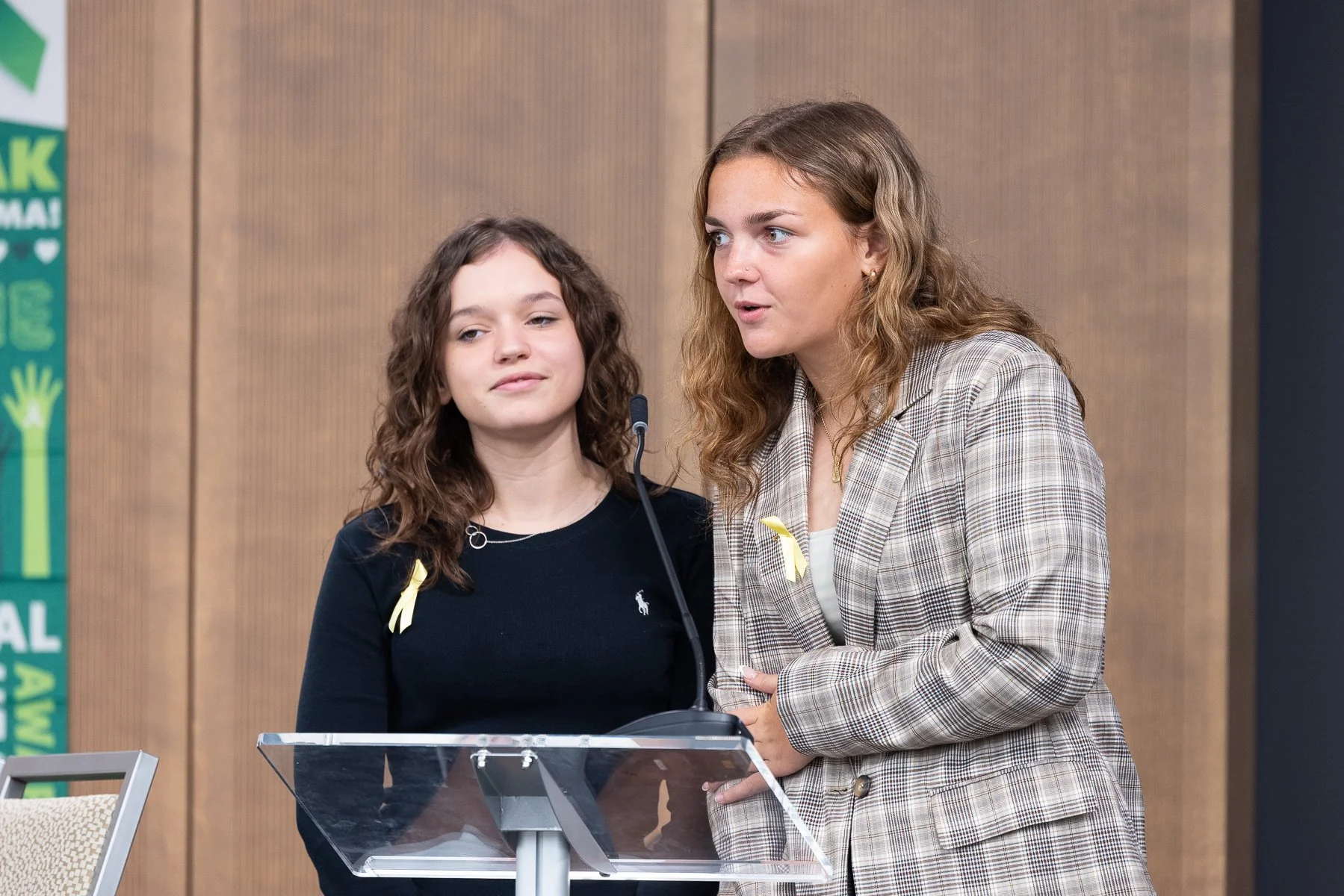 Two women standing at a podium speaking in front of a wooden wall with a green banner on the side.