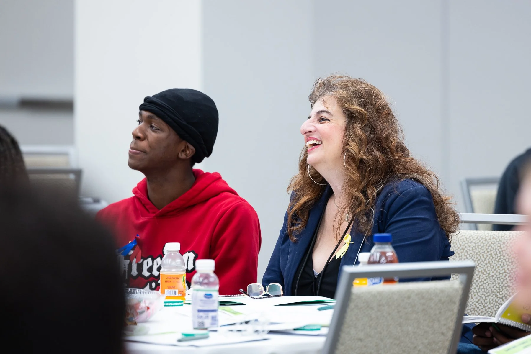 Two women sitting at a table, one in a red hoodie and the other in a navy blazer, smiling and engaging in a discussion during a meeting or seminar, with water bottles and conference materials on the table.